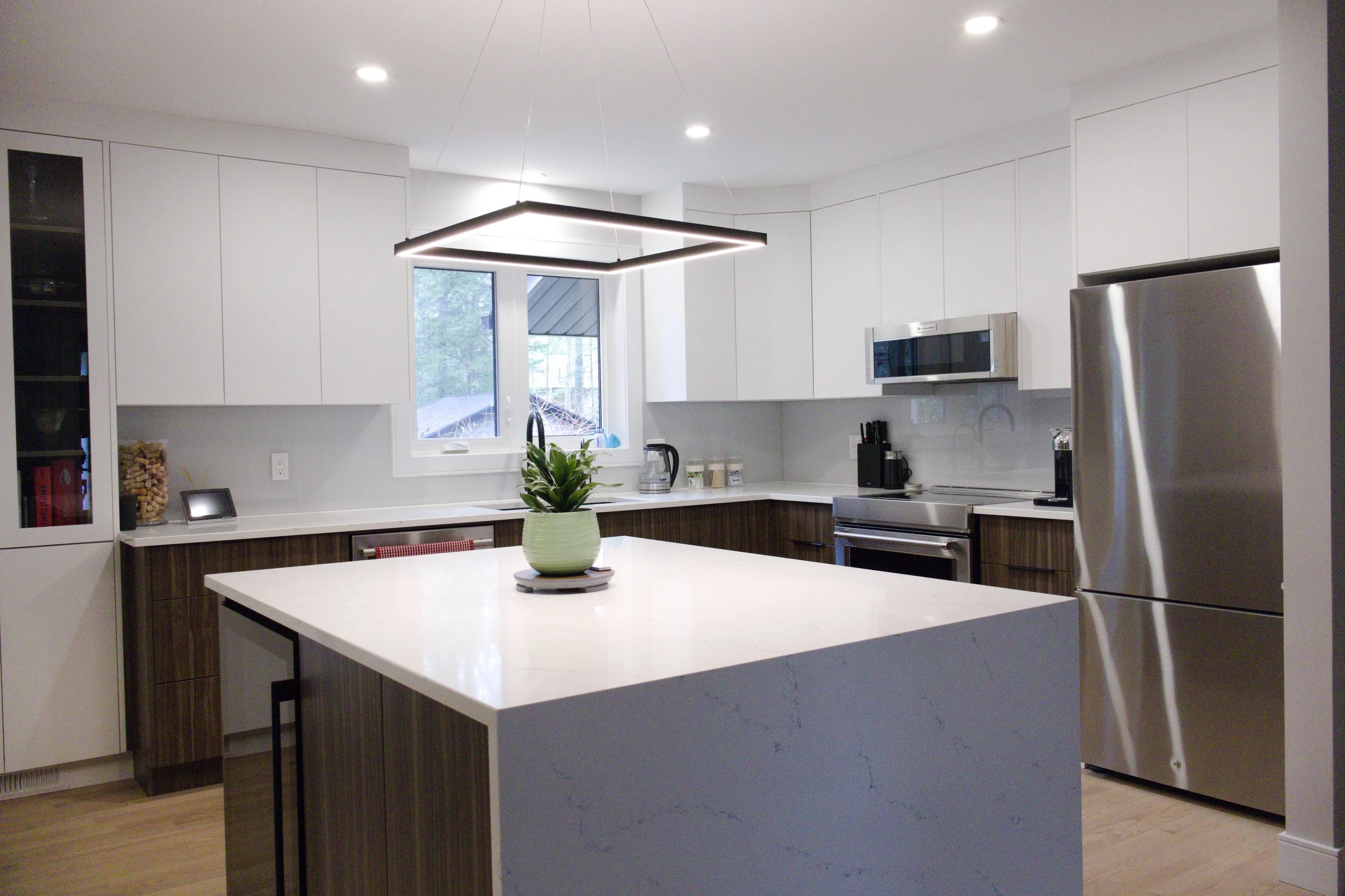 Modern kitchen with white cabinets, stainless steel refrigerator, microwave, black appliances, and a central island with a potted plant, in a well-lit space with a window and light hardwood floors.
