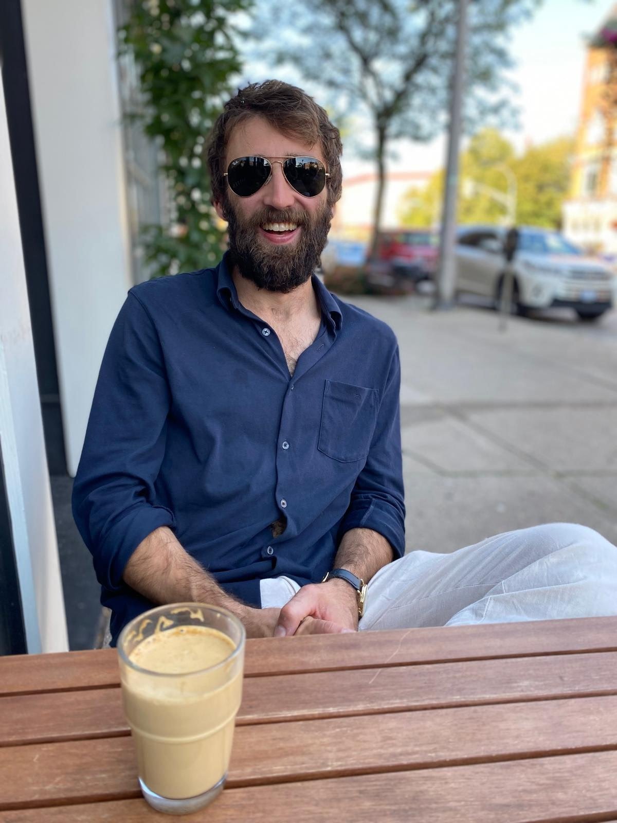 Jaeden, the founder of Foster-Long Custom Builders, smiling at the camera with a beard wearing sunglasses, a blue shirt, and white pants, sitting outside at a wooden table with a pint of beer.