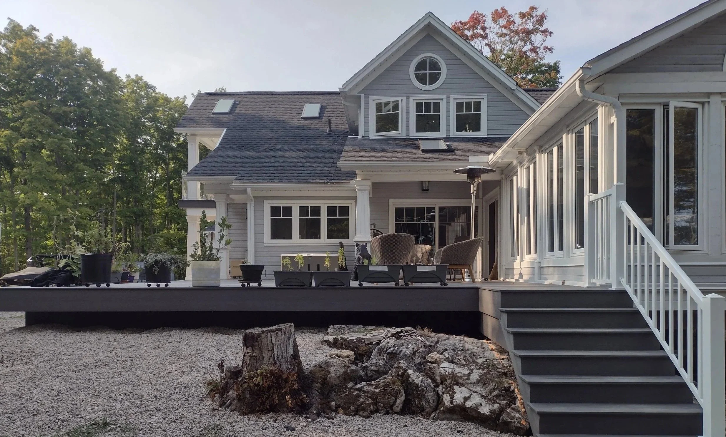Backyard deck of a large house with outdoor furniture, potted plants, and a patio heater, surrounded by trees.