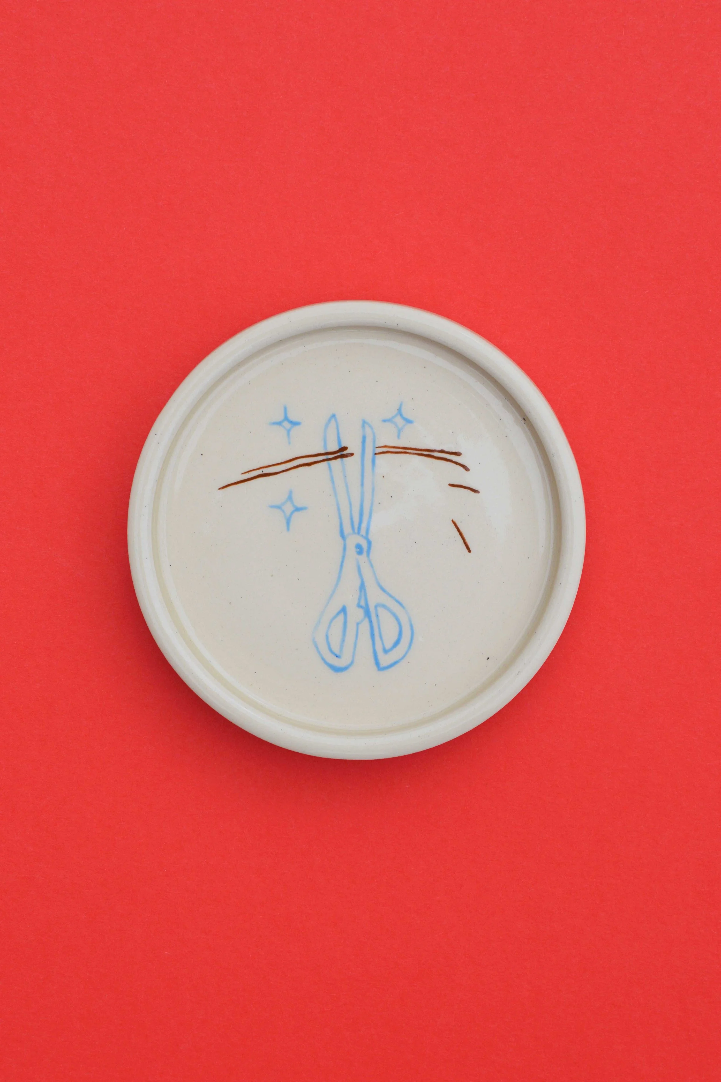 Stoneware ceramic coaster set by artist, Isobel Hill, featuring a pair of blue scissors and pieces of cut brown hair.