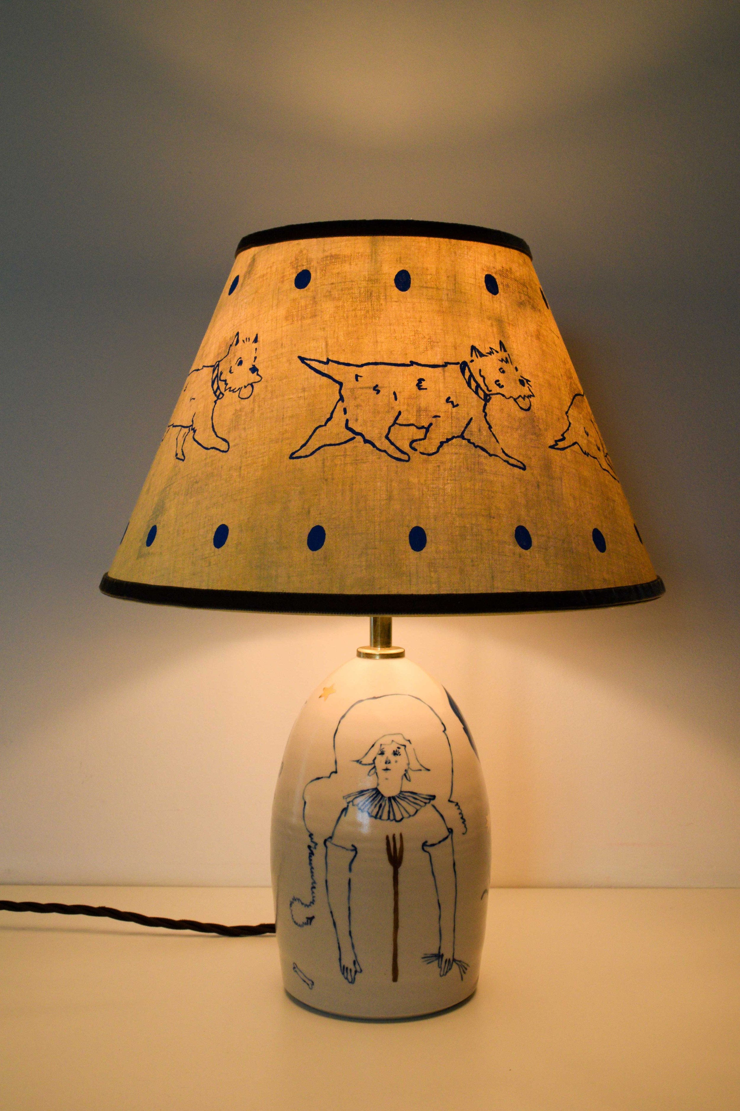 Objects portfolio of artist, Isobel Hill. 2025 Ceramic ‘Trot’ lamp. Illuminated lamp features a handmade linen shade with running Westie and an underglaze and gold lustre design of a figure with a pitchfork. 