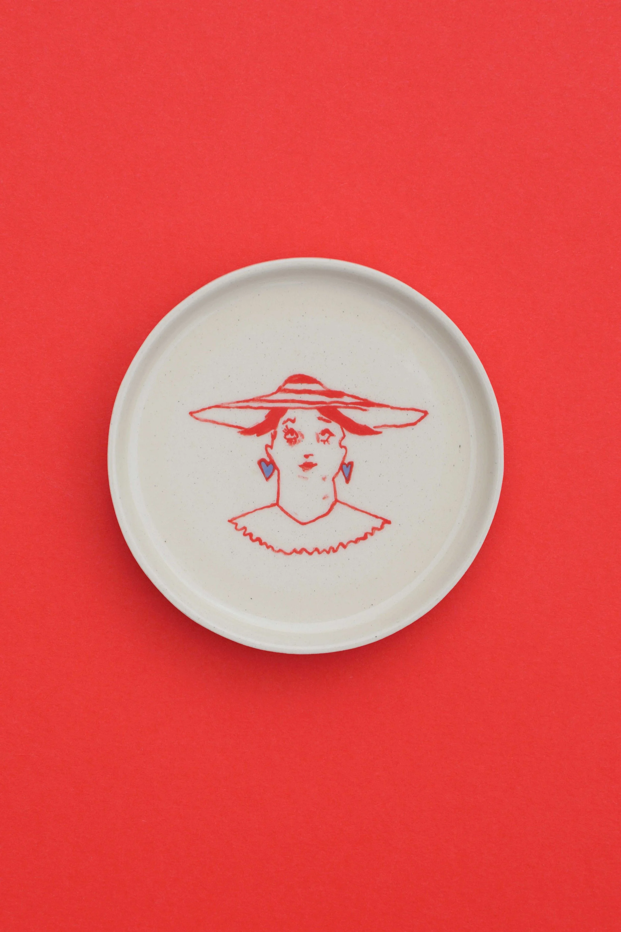 Stoneware ceramic coaster set by artist, Isobel Hill, featuring a person in a red brimmed stripe hat and periwinkle heart earrings.