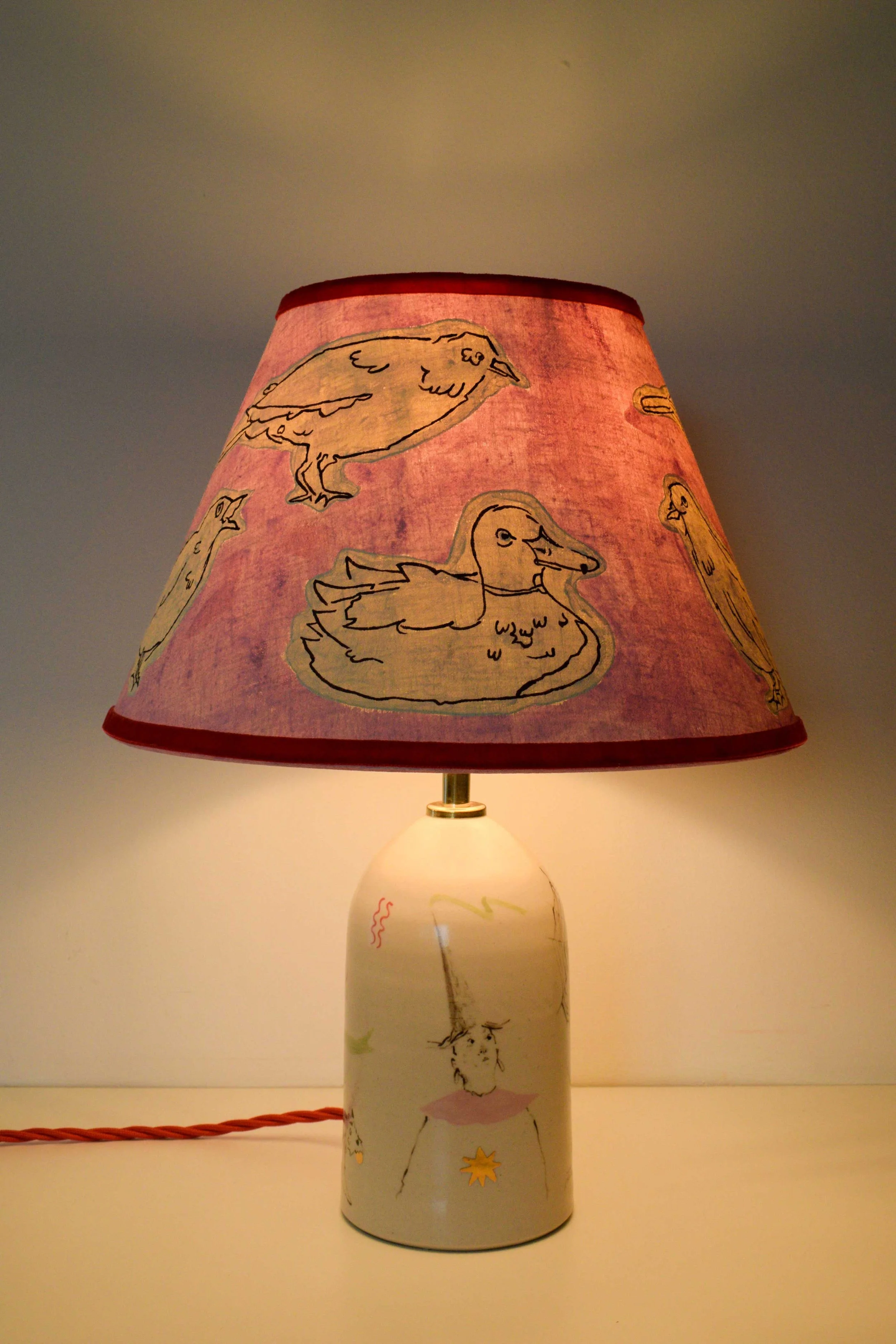 Handmade ceramic lamp base by artist, Isobel Hill, with painted pink duck linen lampshade and illustrative figurative design. Lamp is on.