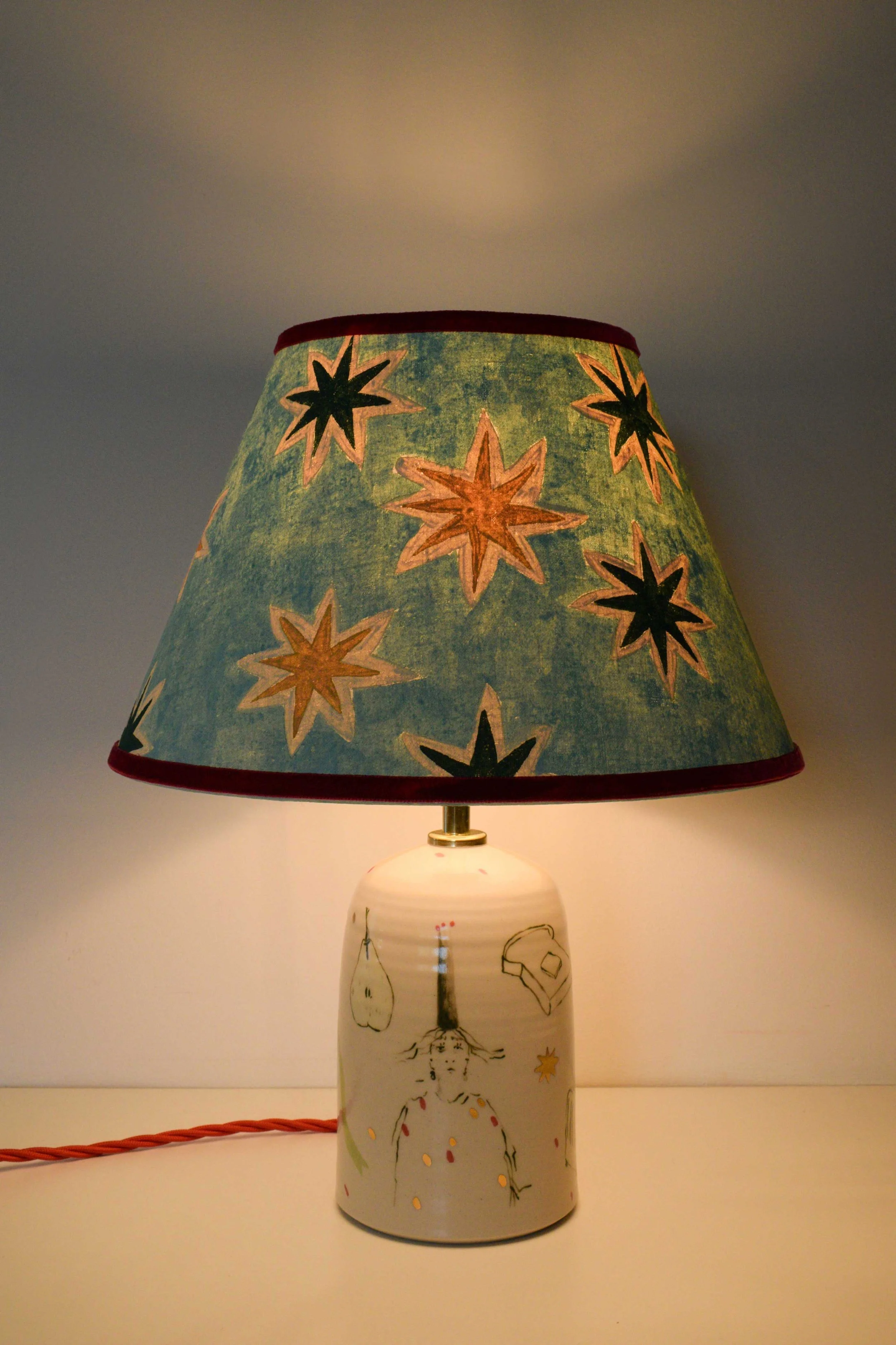 Handmade ceramic lamp base by artist, Isobel Hill, with painted stars linen lampshade and illustrative figurative design. Lamp is on.