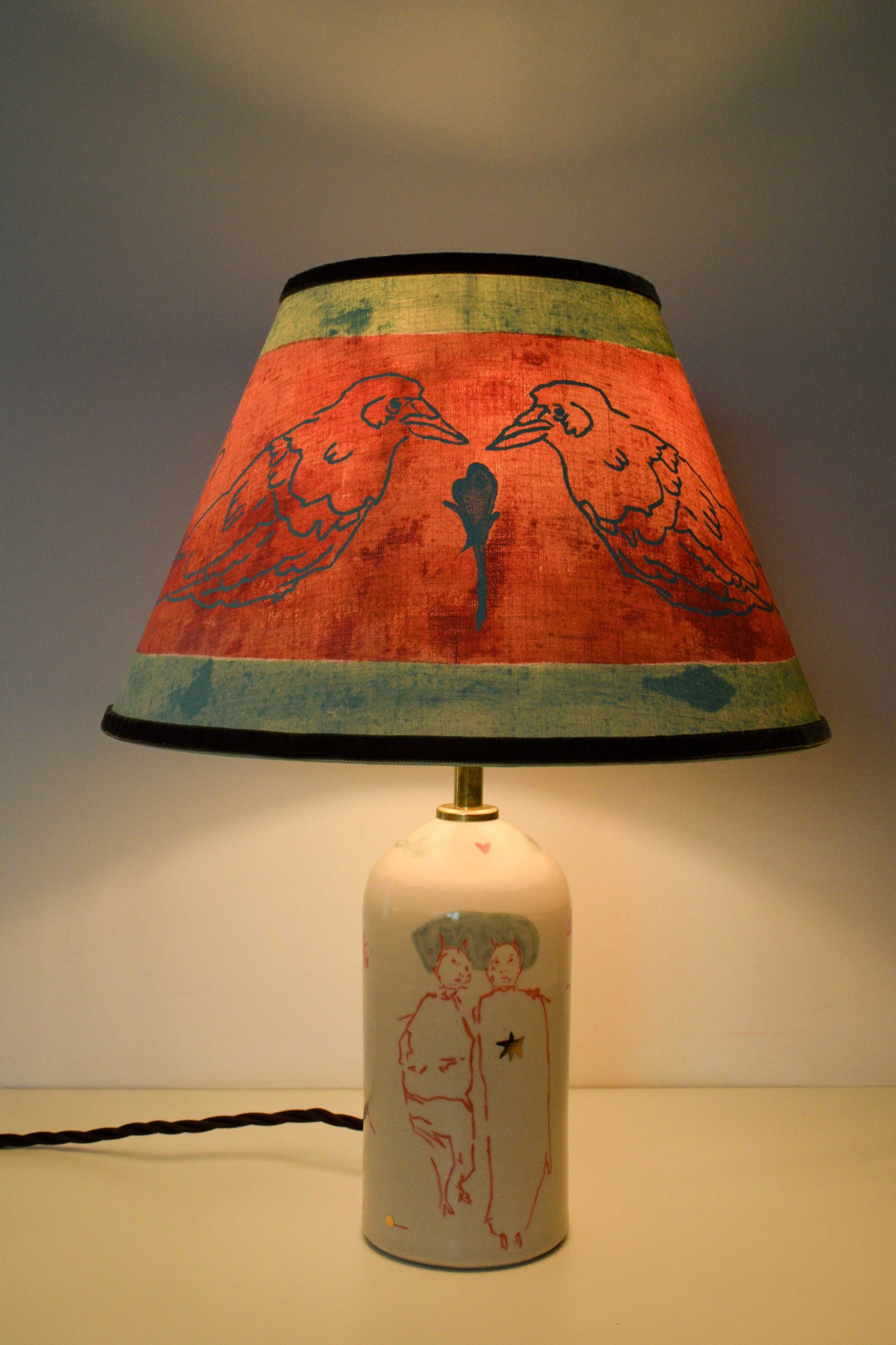 Objects portfolio of artist, Isobel Hill. 2025 Ceramic ‘Crow’ lamp. Illuminated lamp features a handmade linen shade with mirrored crows and an underglaze and gold lustre design of two figures laying down with a gold star.