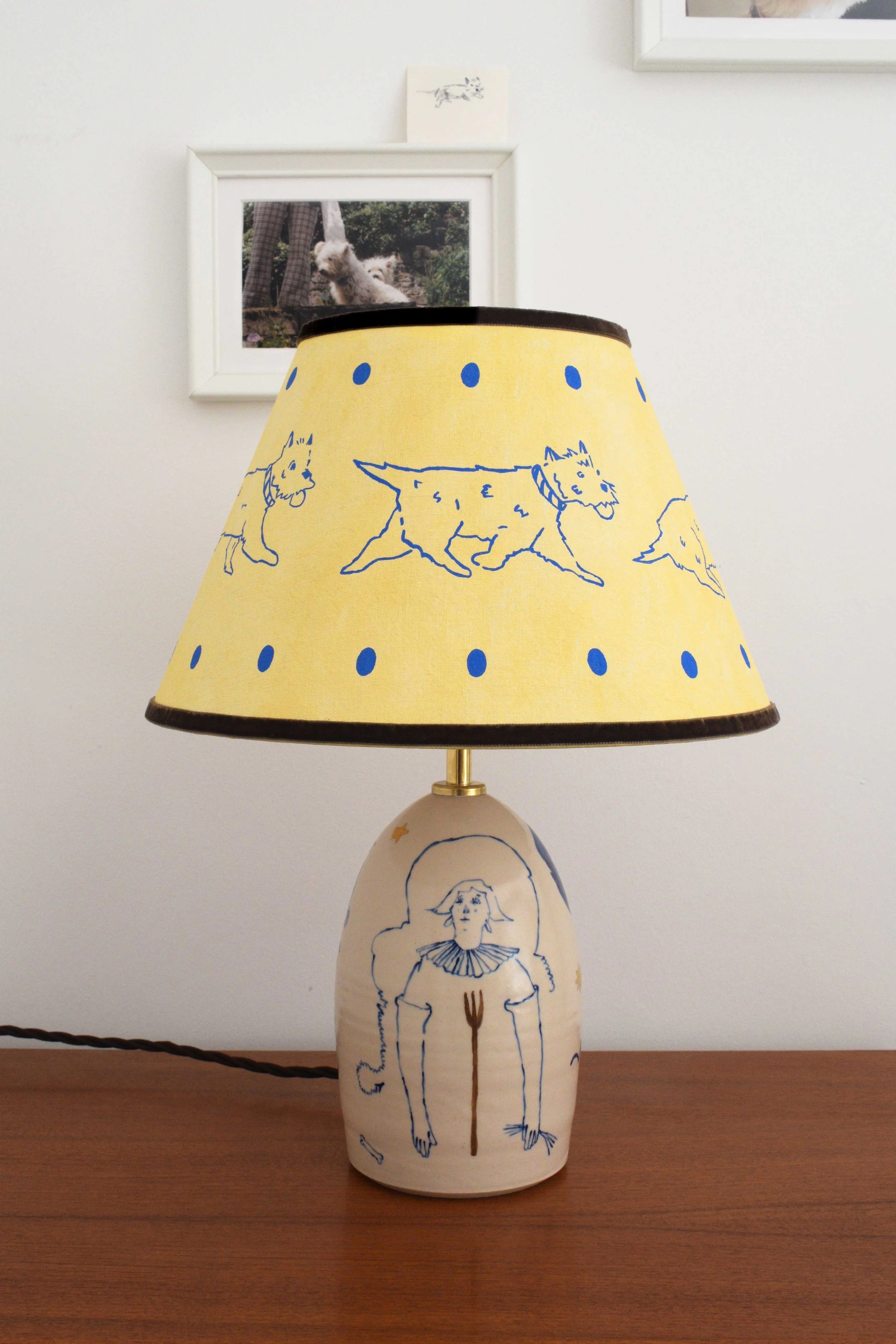 Objects portfolio of artist, Isobel Hill. 2025 Ceramic ‘Trot’ lamp. Lamp features a handmade linen shade with running Westie and an underglaze and gold lustre design of a figure with a pitchfork. 