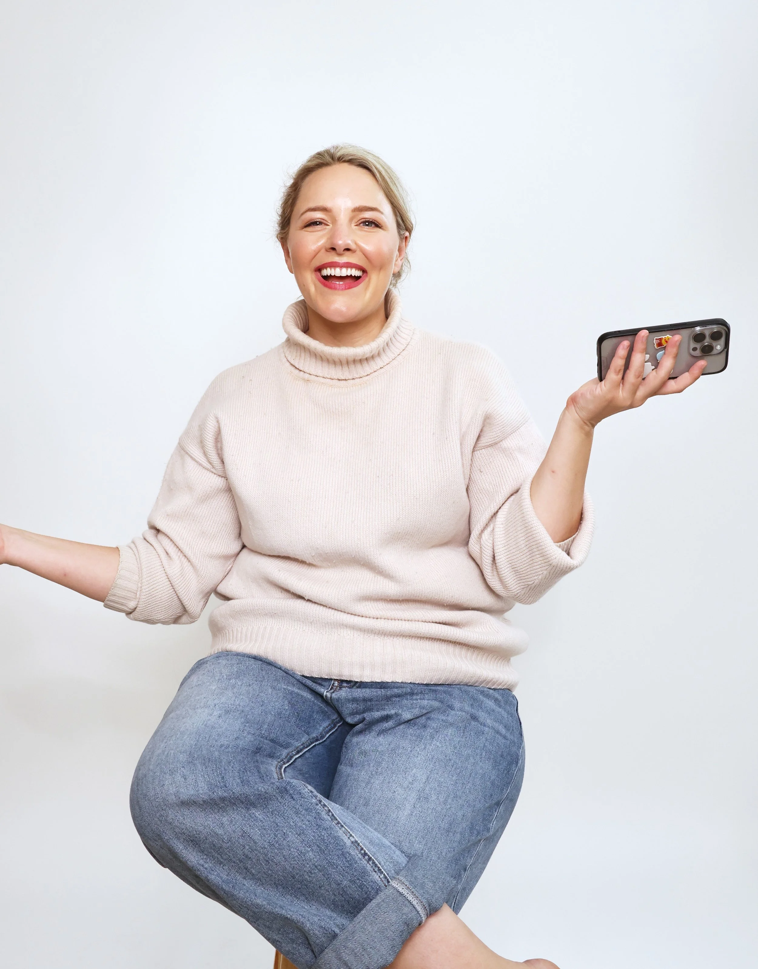 Ettie is a white woman with dark blonde hair. She is wearing a cozy wool turtleneck jumper with jeans, and is holding her phone in her hand. Ettie has a big smile on her face.