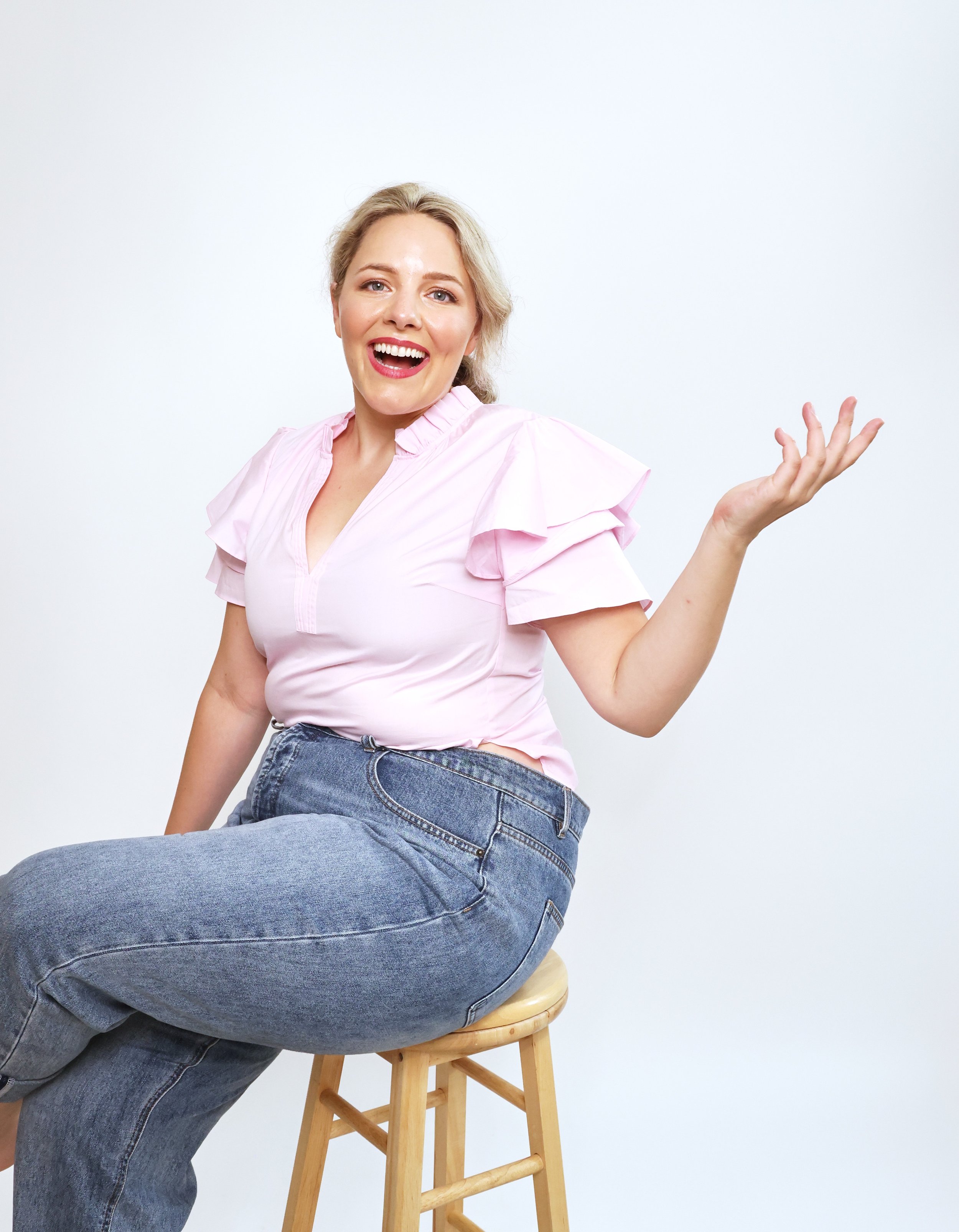 Ettie is a white woman with dark blonde hair. She is sat on a wooden stool and smiling joyfully to the camera.