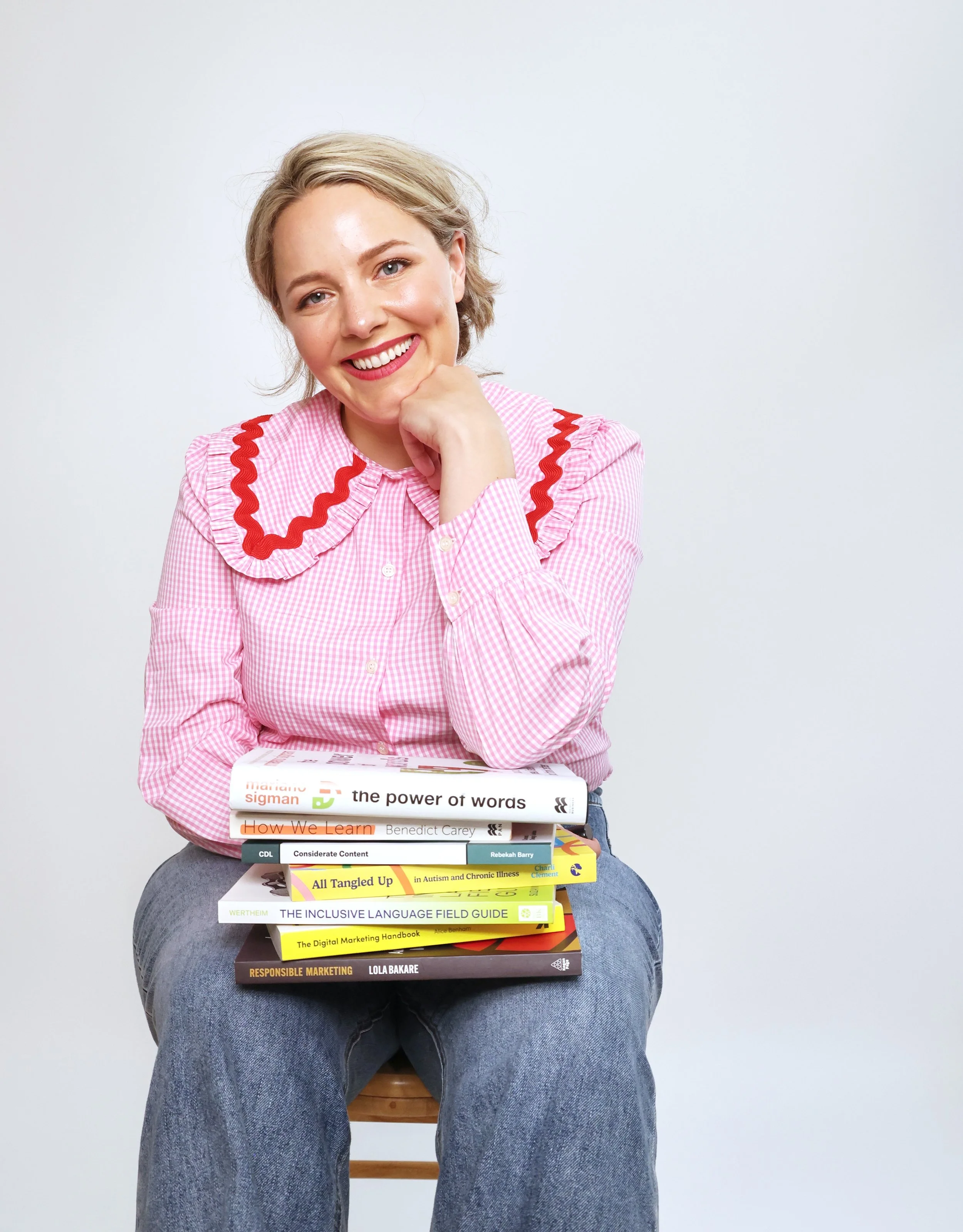 Ettie is a white woman with dark blonde hair. She is sat down with a pile of books about inclusive and accessible communication on her lap.