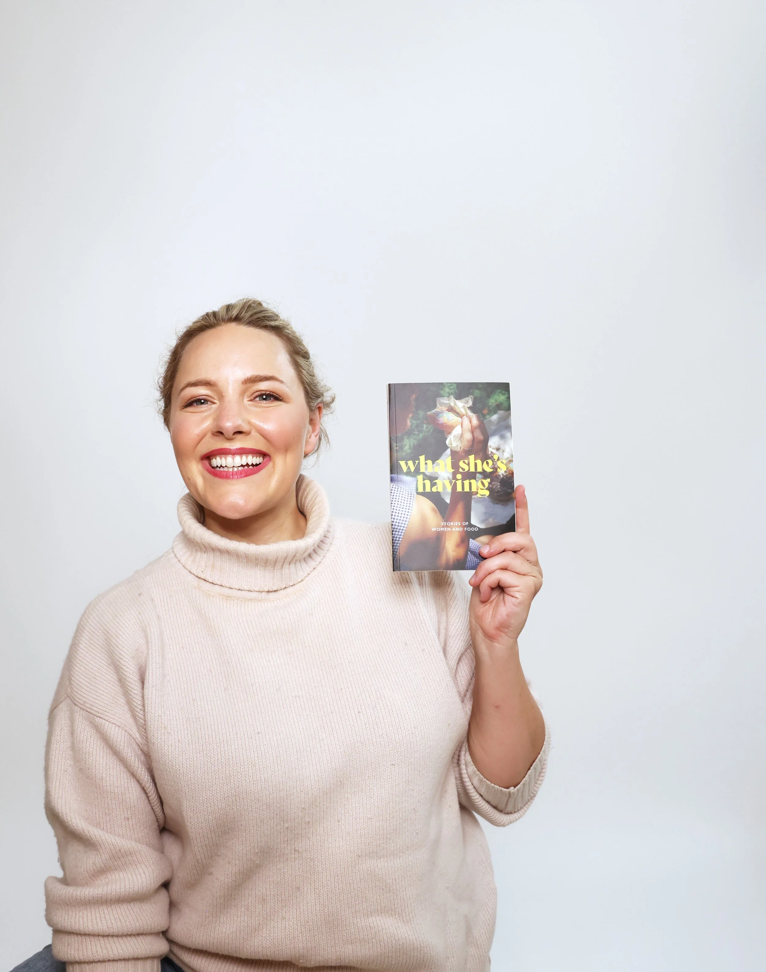 Ettie is a white woman with dark blonde hair. She is wearing a cozy turtleneck jumper and holding up the book “What She’s Having: Stories of Women and Food”, which features her short story “Feast or Foe”.