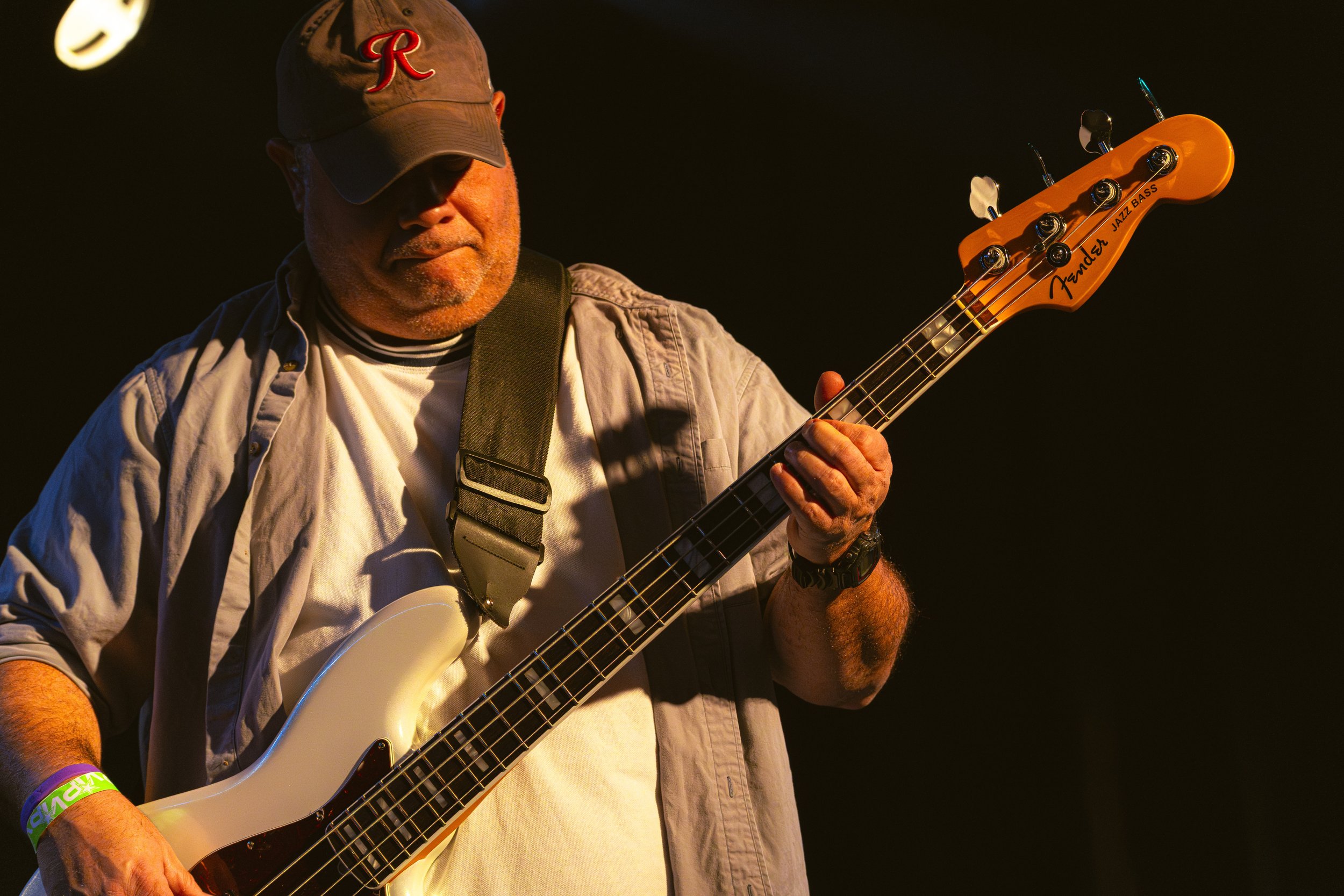 Man playing a white electric bass guitar on stage, wearing a beige jacket, cap, and wristbands, with a dark background.