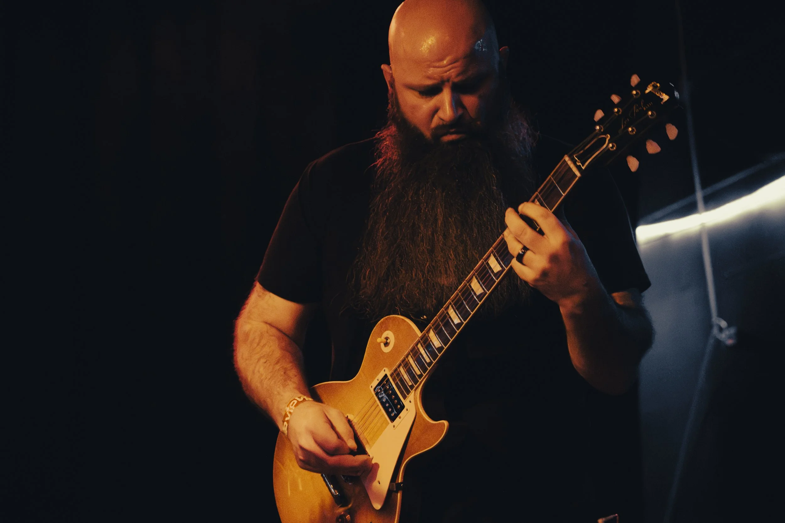A man with a beard playing an electric guitar on stage in a dark setting.