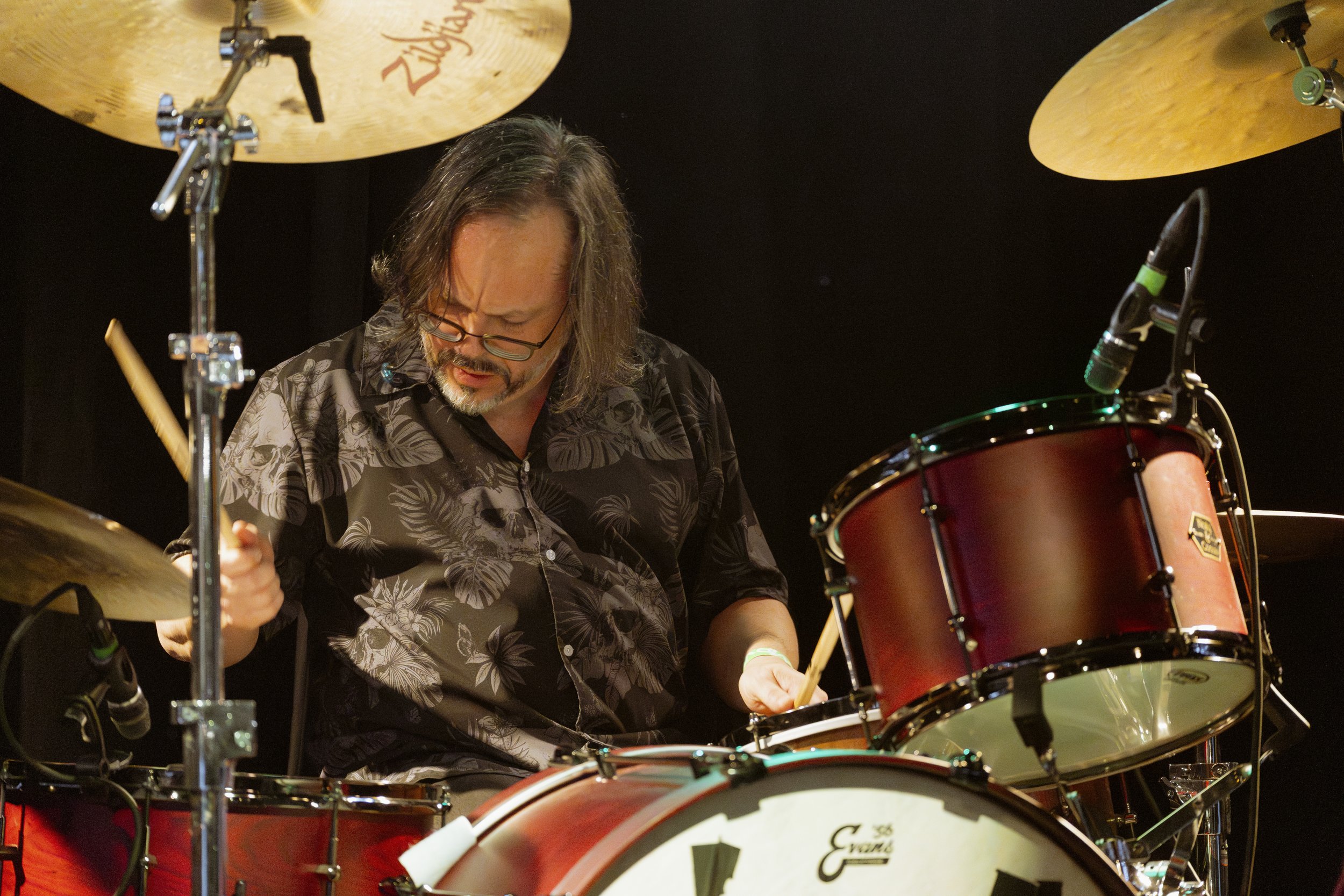 A man with long hair and glasses playing a drum set during a performance, with cymbals and microphones visible against a dark background.