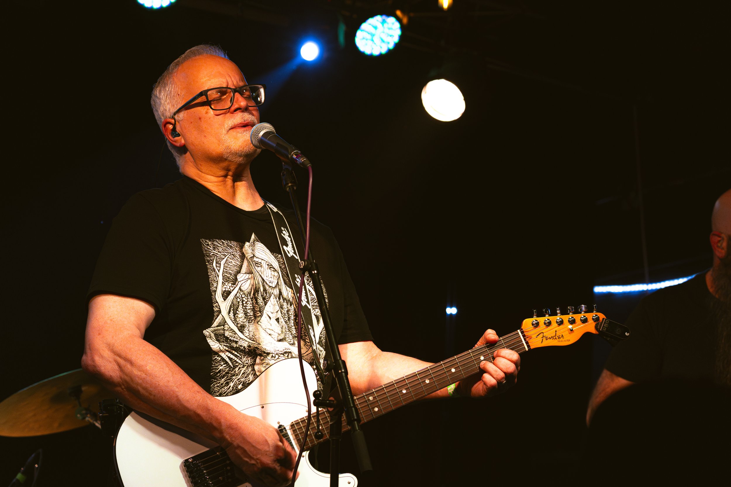 An older man with glasses and gray hair is singing into a microphone while playing a white Fender electric guitar on stage under colorful stage lights.