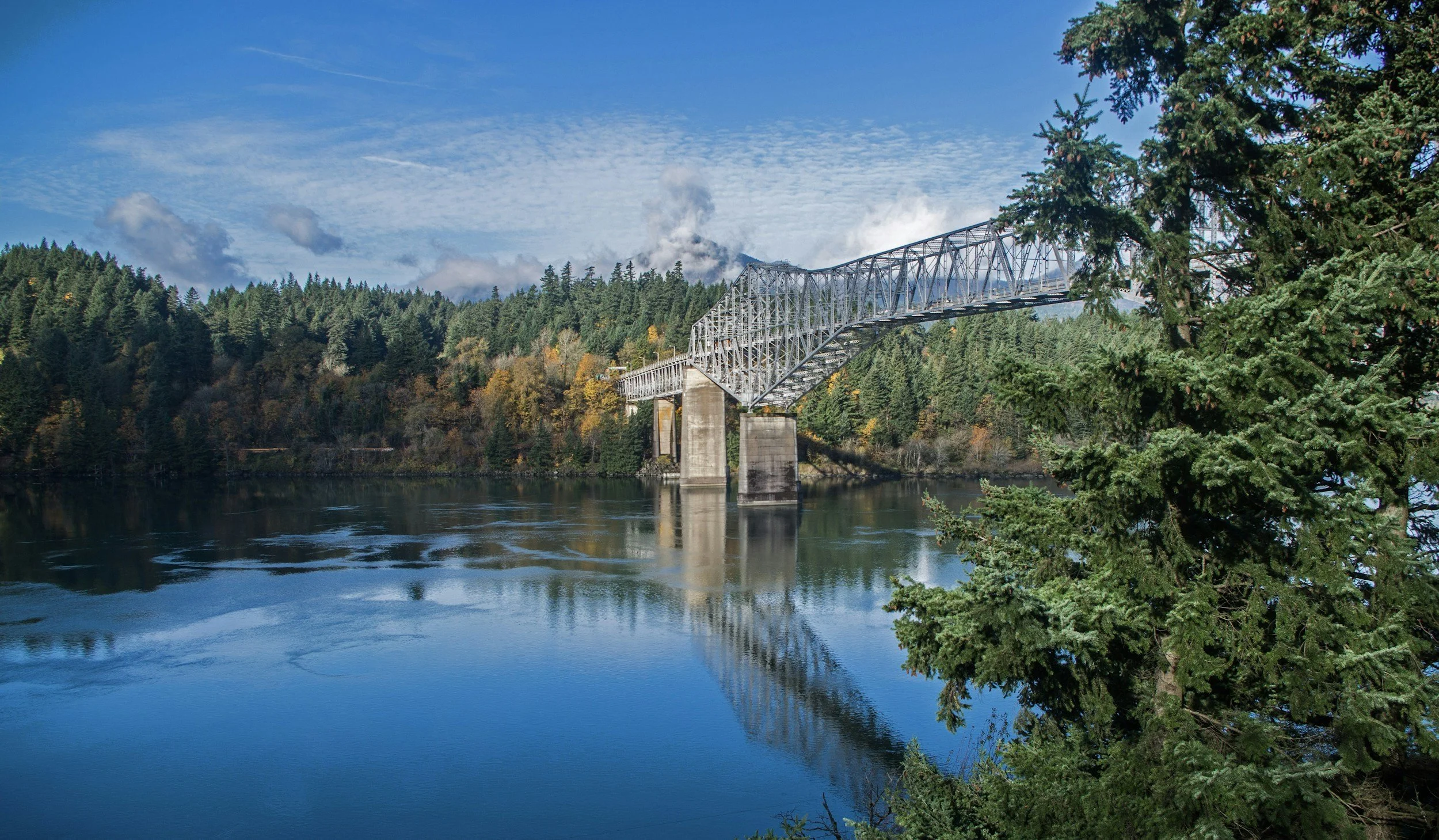 Photo of the Bridge of the Gods facing north from near river-level in the fall. The water is calm with a rippling reflection of blue sky and a few clouds.