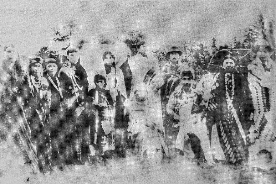 Black and white family photo of Warcomac and Miller family circa 1898. A group of adults and children of varying ages stand, looking at the camera,  posed in traditional regalia.