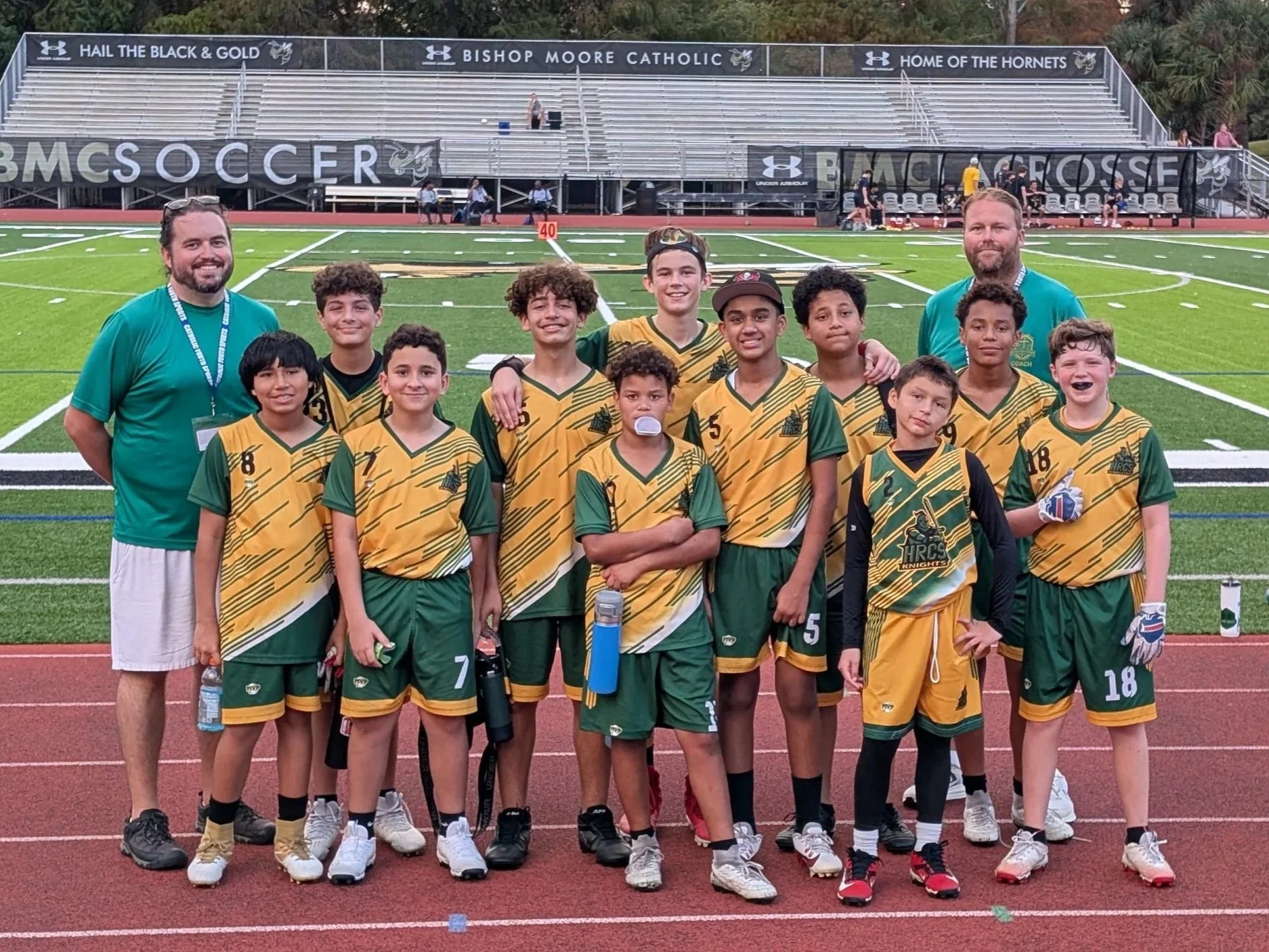 A youth basketball team with coaches standing on a sports field. The players are wearing yellow and green uniforms with numbers, and the coaches are in teal shirts. The group is posing for the photo with a stadium and bleachers in the background.