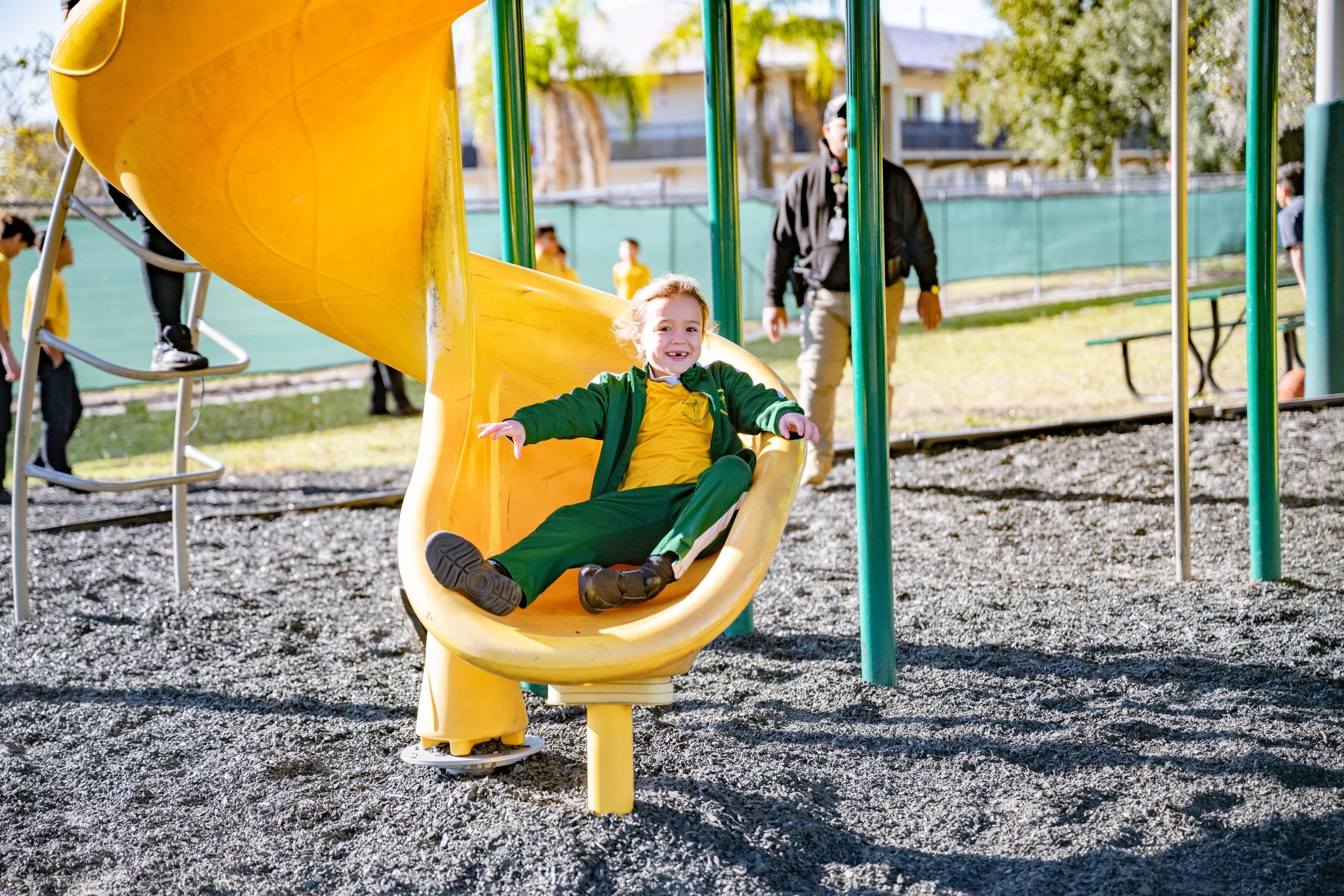 Child smiling and sliding down a yellow playground slide in a park with green trees and a fence in the background.