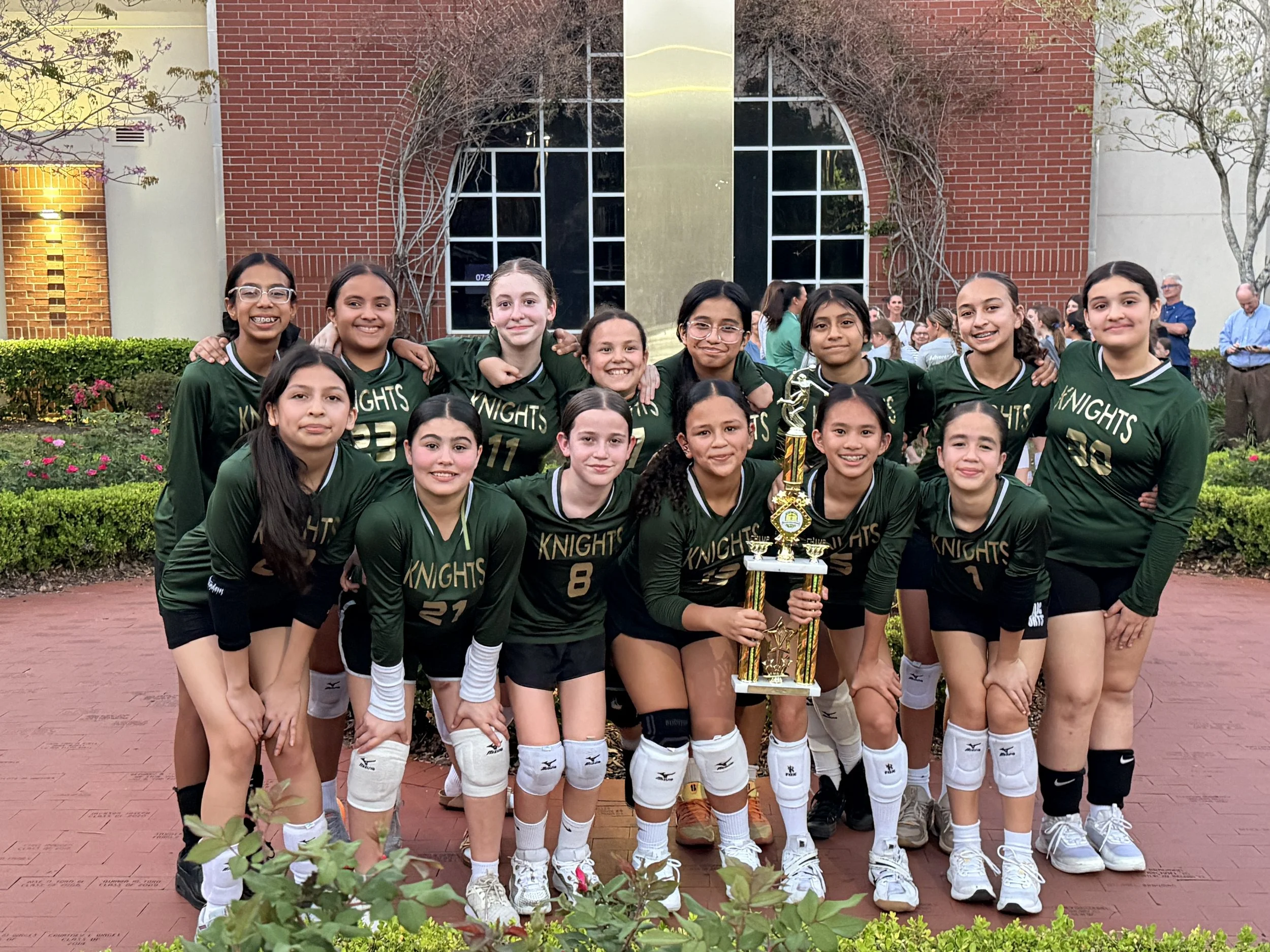 A group of young girls in green sports jerseys with the word ’Knights’ and numbers, posing outdoors on a red-brick pathway in front of a building. They are smiling, with some holding a trophy. The background features a brick building with a large window, trees, and other people.