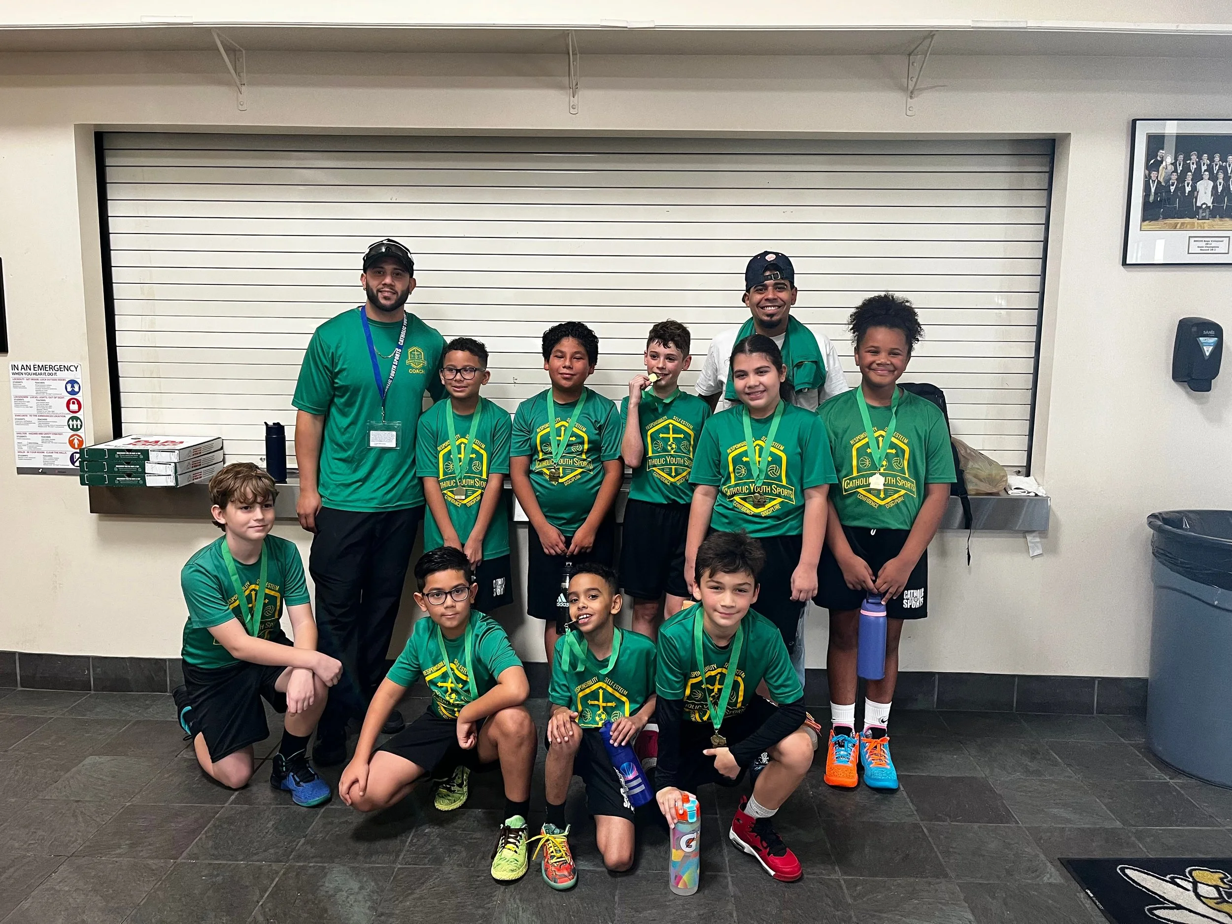 Group of children and two coaches wearing green shirts with sports medals, posing for a photo indoors in front of a metal shutter.