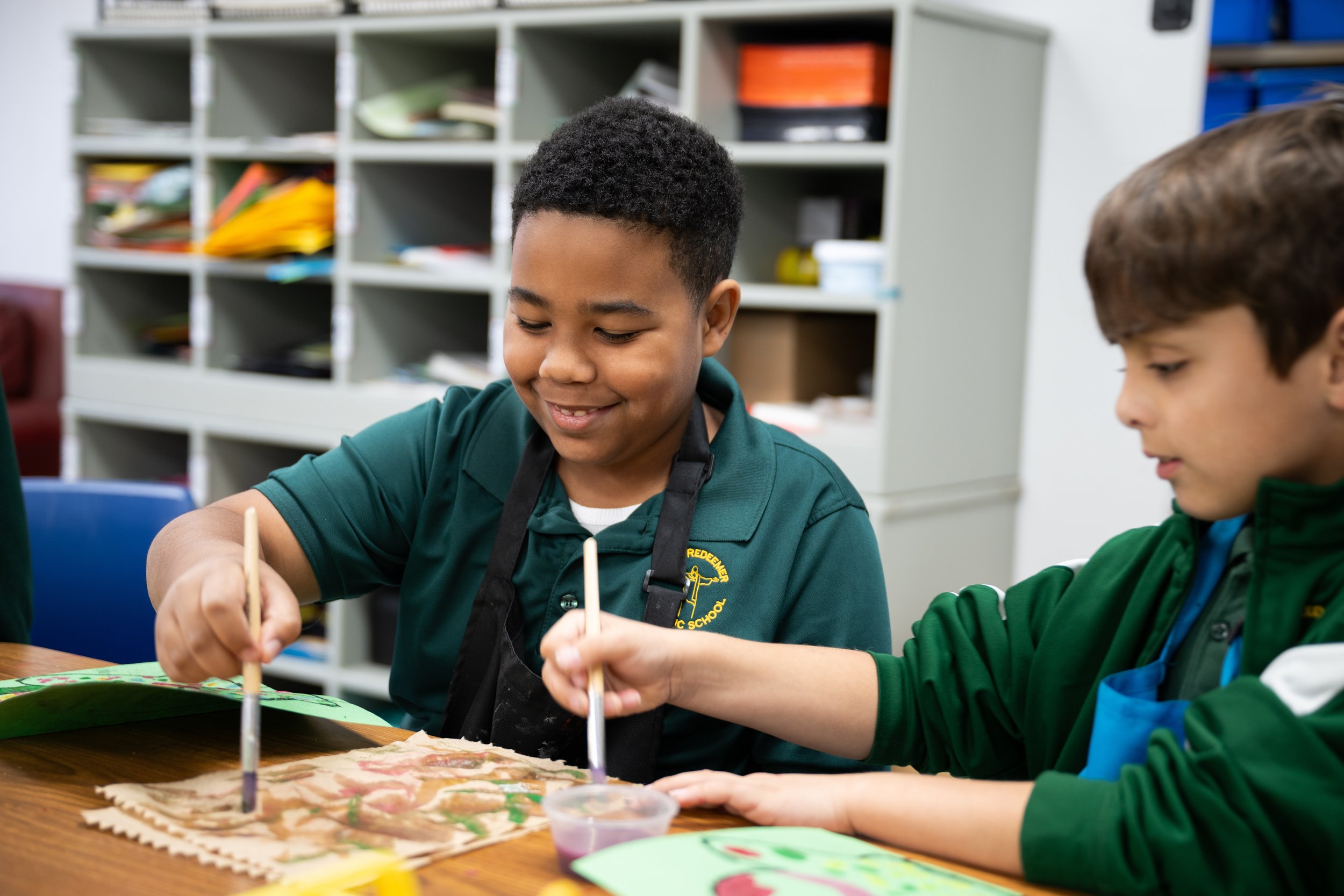 Two young boys, dressed in green uniforms, sitting at a table and engaging in an arts and crafts activity. They are smiling and working with wooden sticks, with art supplies spread on the table.