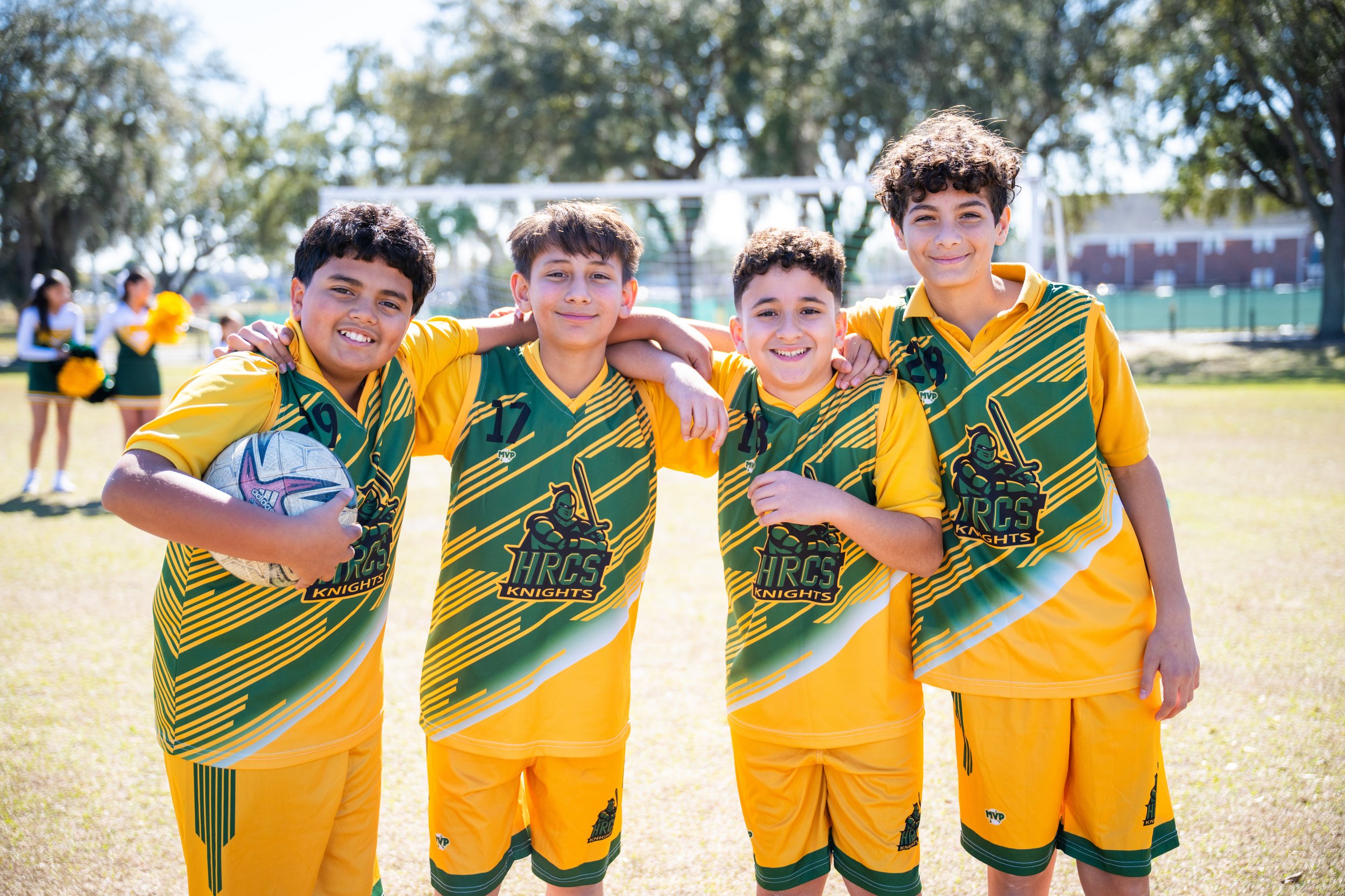 Four boys in yellow and green sports uniforms standing close together on a grassy field with trees, smiling for a photo, with cheerleaders and a soccer goal in the background.