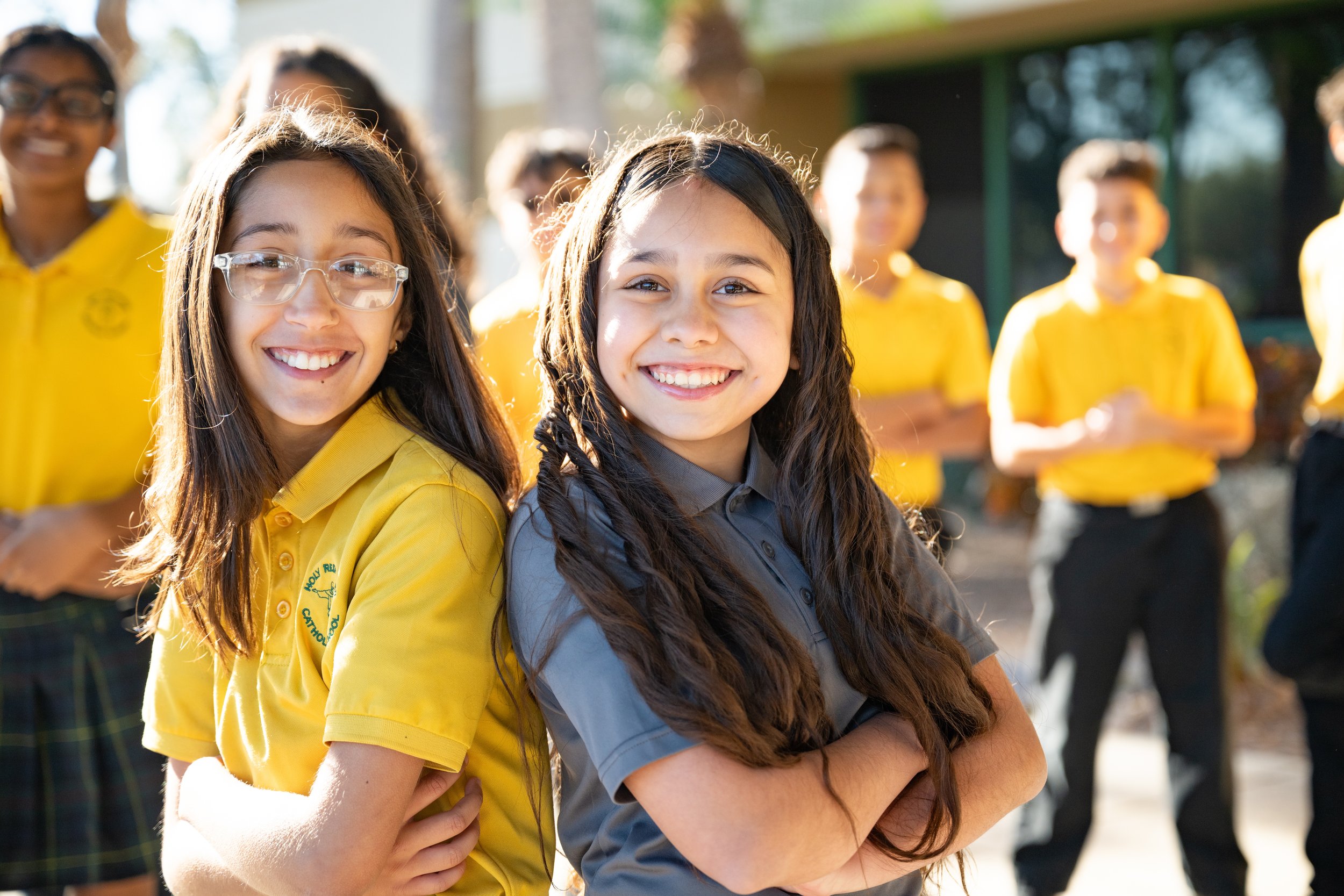 Smiling children with crossed arms outdoors, wearing yellow and gray uniforms, standing in a schoolyard.