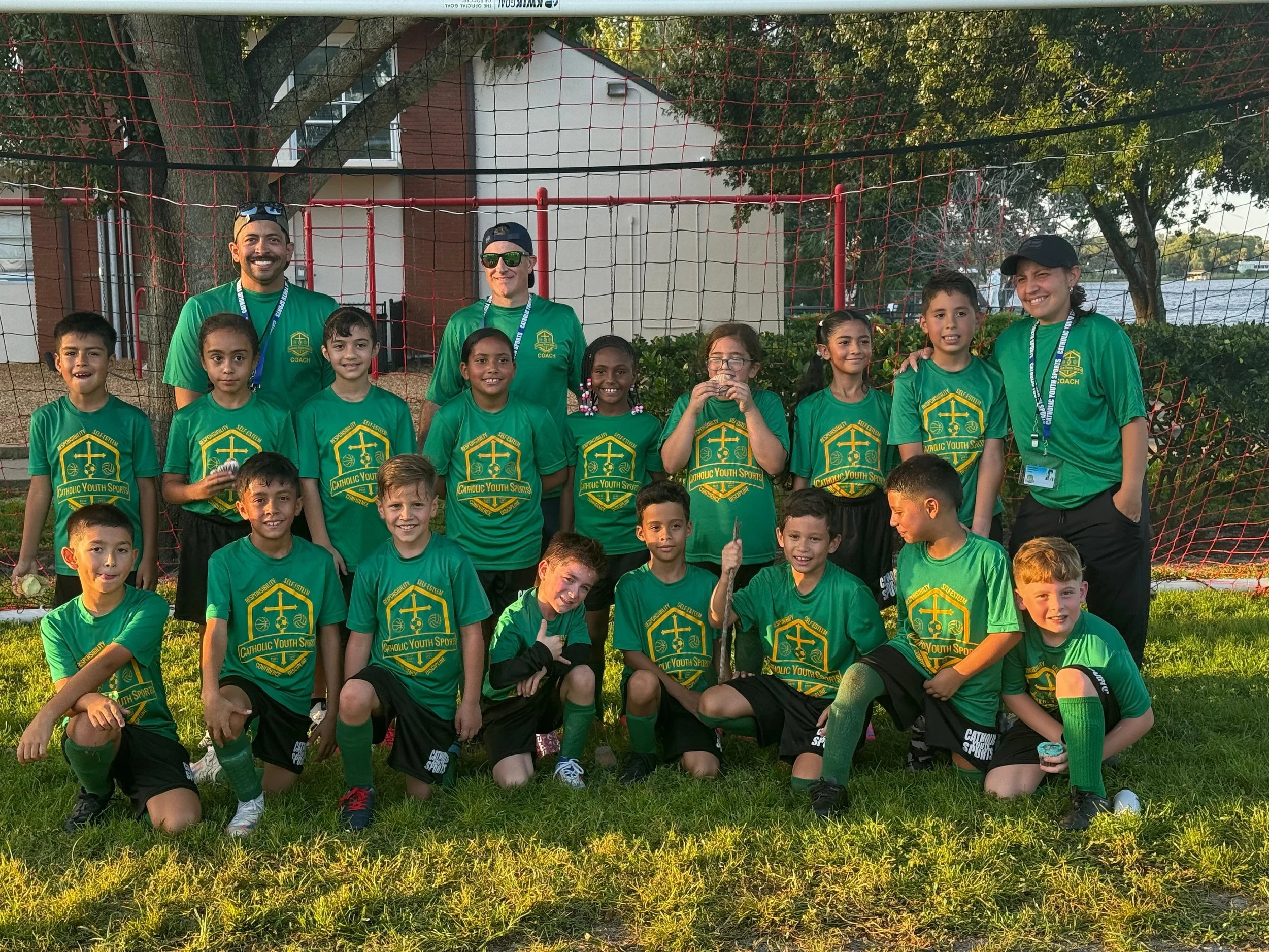 A group of children and adults in green sports shirts posing outdoors in front of a red safety net, with trees, a building, and water in the background.