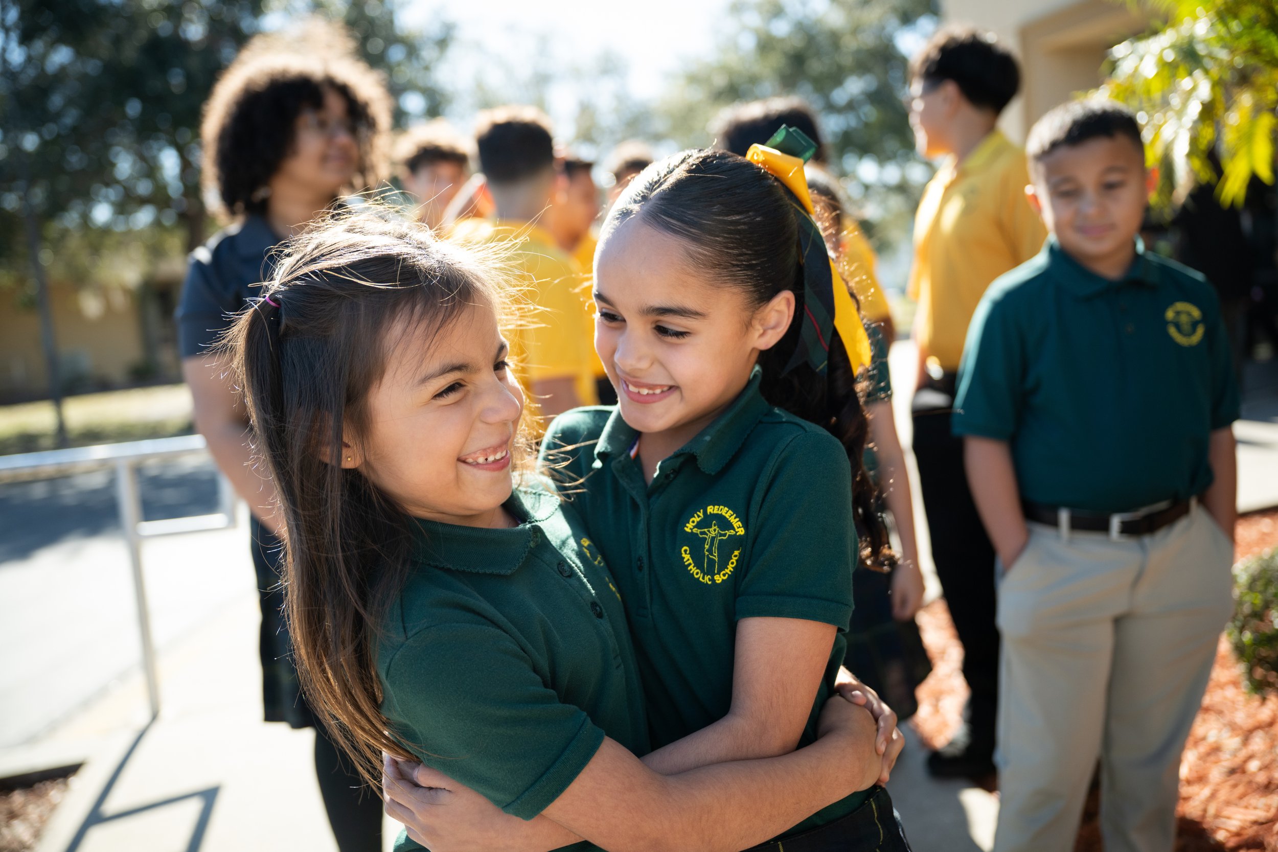 Two young girls hugging outdoors on a sunny day with a group of children and adults in the background.