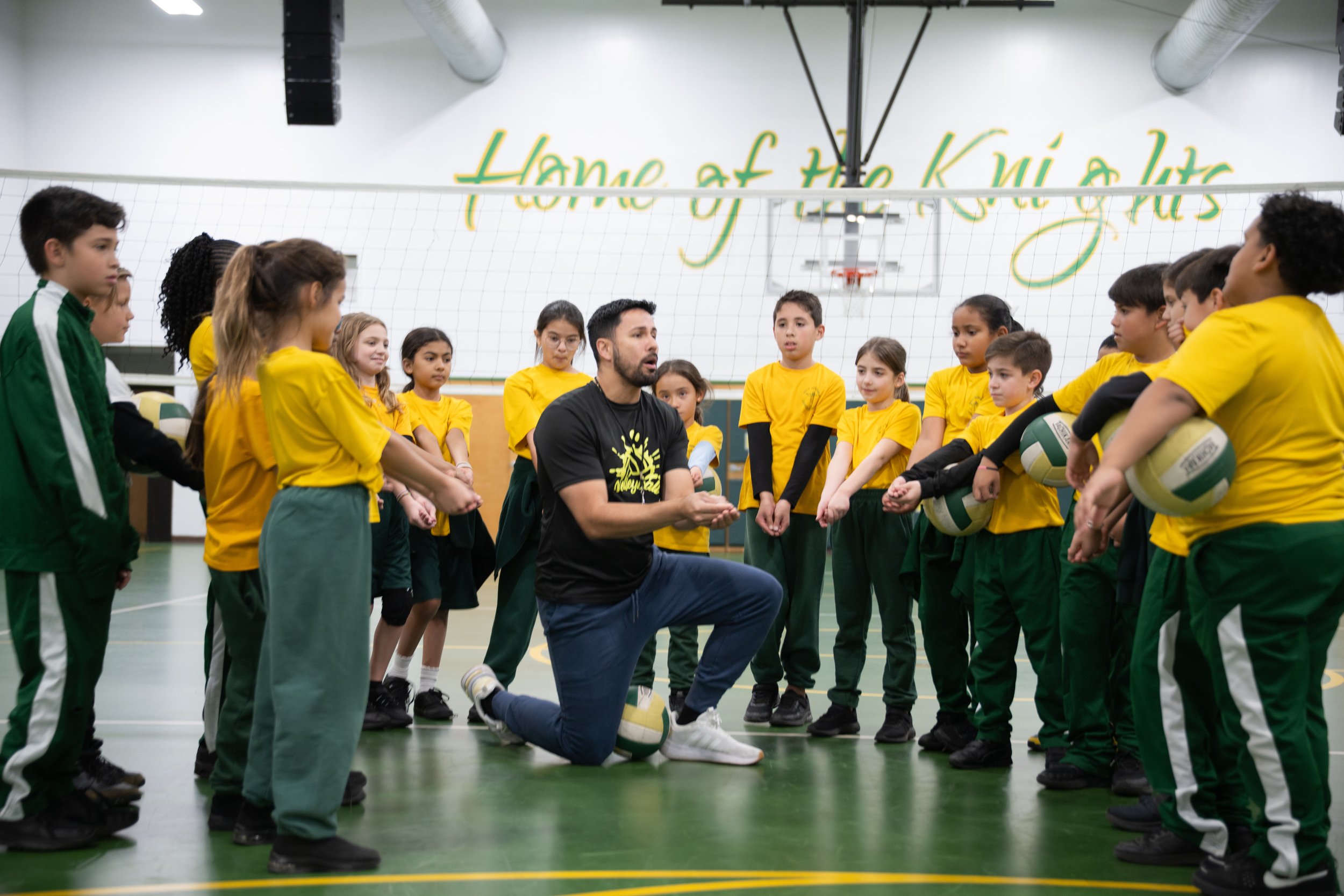A group of children in yellow and green sports uniforms listening to their coach, who is kneeling and speaking to them in a gymnasium with a volleyball net and painted motivational words on the wall.