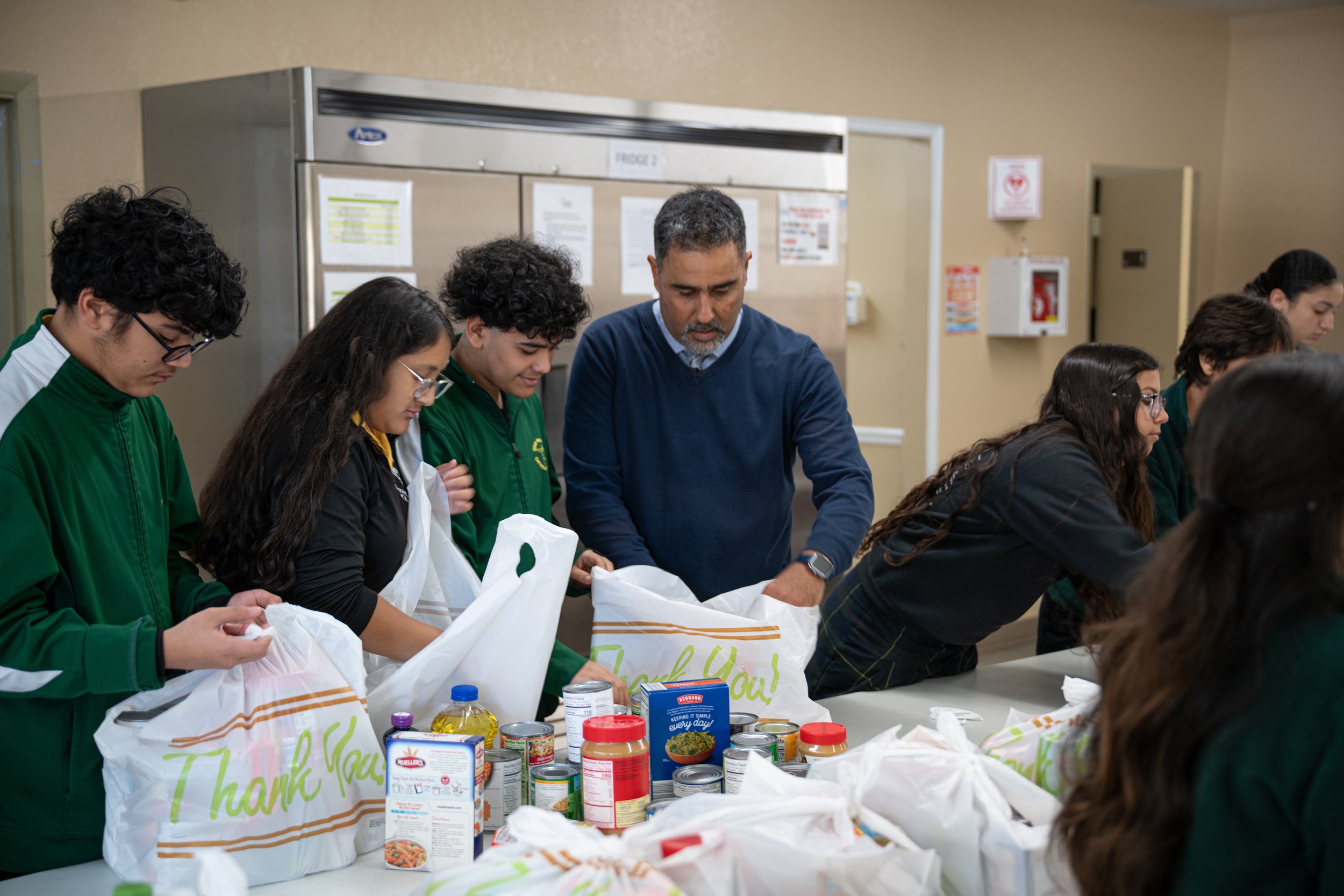 People, including a man and several young individuals, are standing at a table sorting groceries, including canned goods and other food items, in a room with beige walls and informational posters in the background.