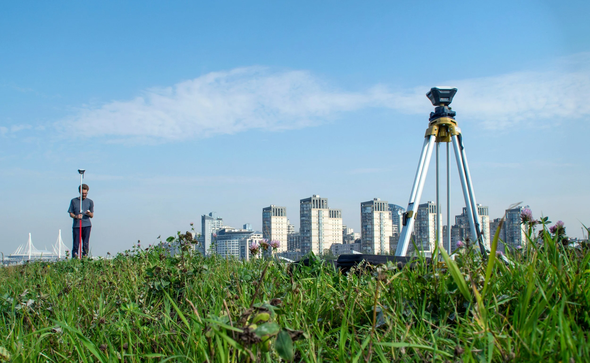 A person using surveying equipment outdoors on a grassy field with tall buildings in the background, a tripod in the foreground, and a bright blue sky.