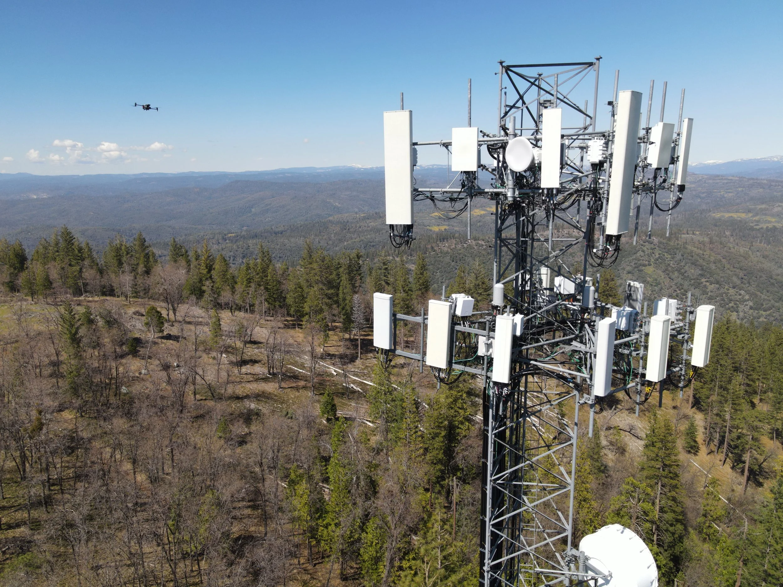 A cellphone tower with multiple antennas and dishes stands in a forested mountainous area under a clear blue sky. A drone is flying nearby in the sky.