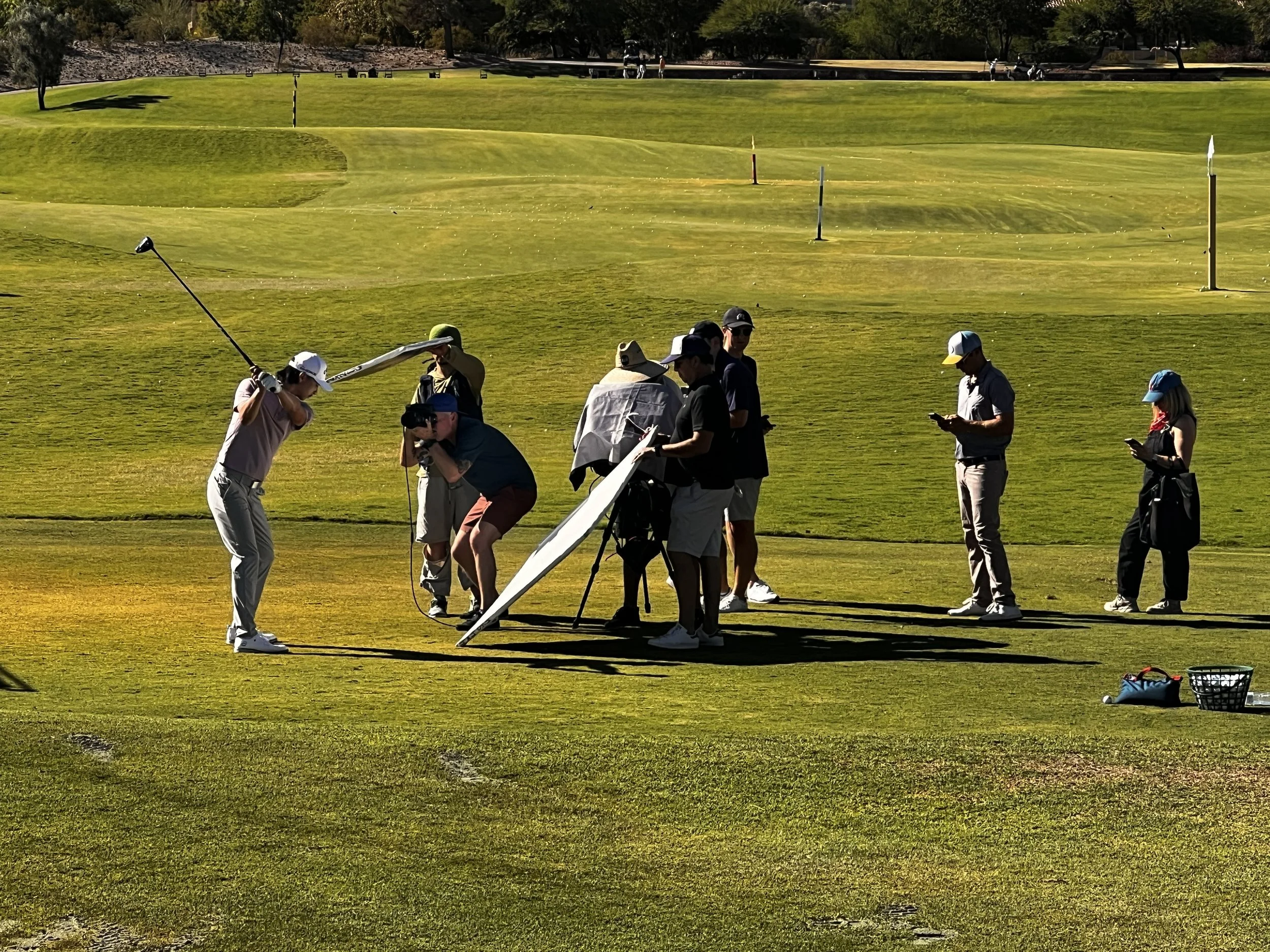 Group of people practicing golf on a golf course, with some preparing to take a shot and others observing or using their mobile phones under sunny weather.