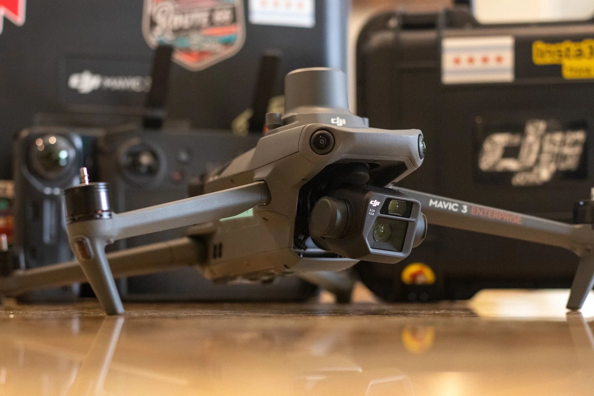 A DJI Mavic 3 Enterprise drone resting on a wooden floor, with its camera and sensors visible, surrounded by cases and remote controls in the background.