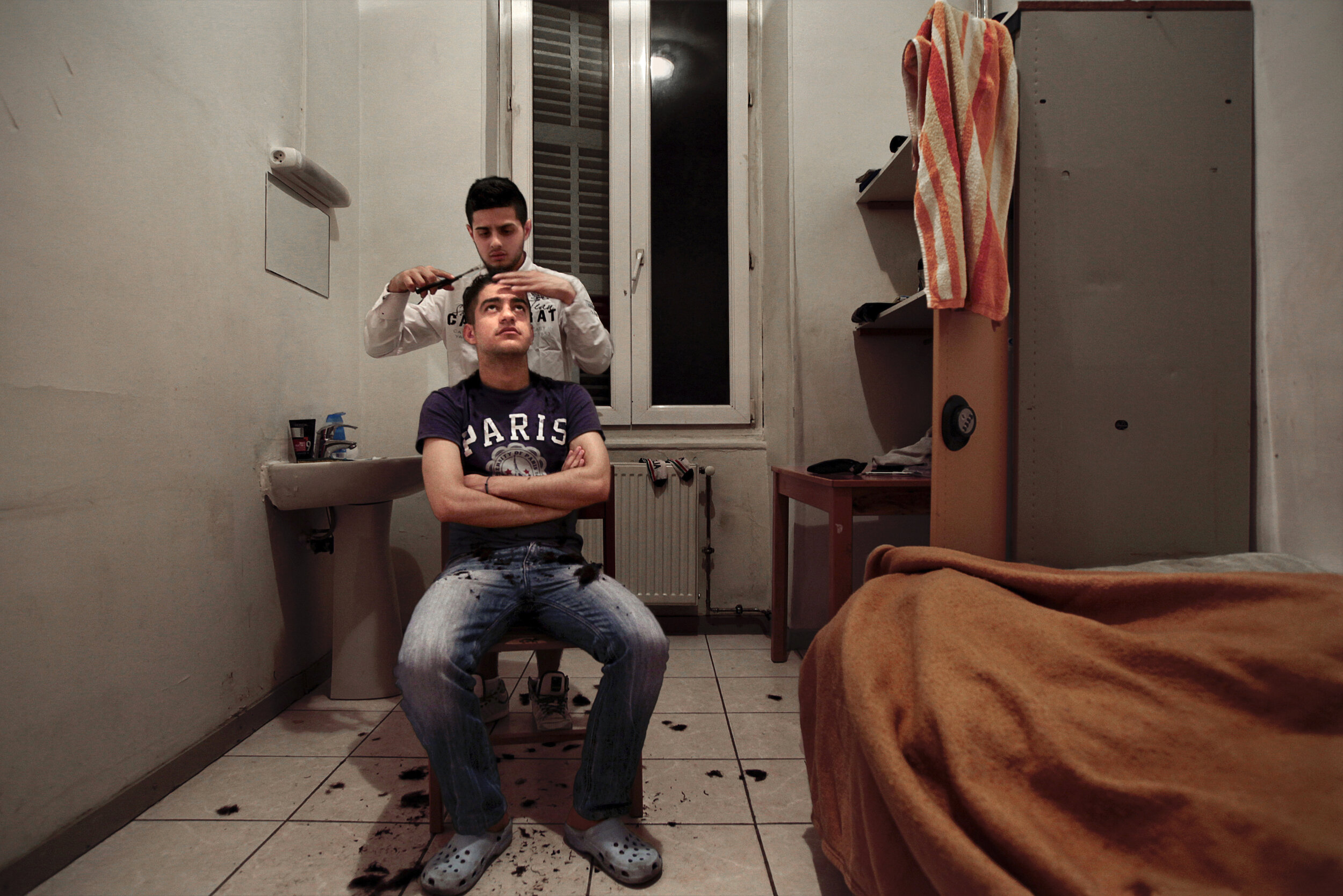   France. Marseille. 2014. Inside his small room in a shelter for homeless young men, Daniel (standing), 19, gives a haircut to his friend, Mohamed Chérif Spiga. Daniel dreams of becoming a hairstylist and provides his services free of cost to all th
