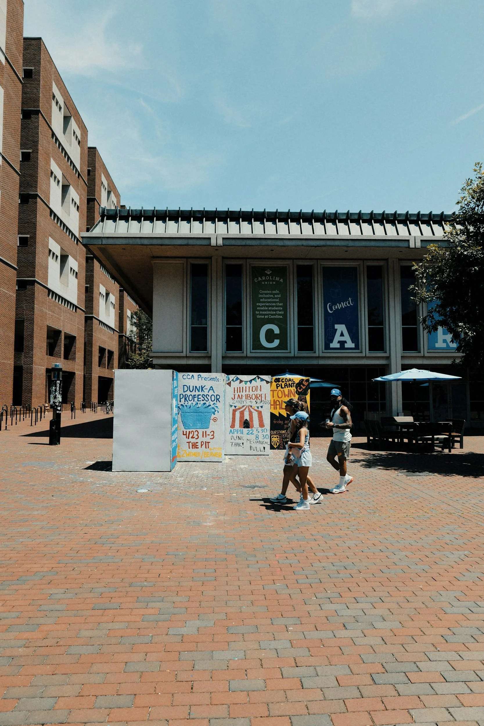 A brick-paved outdoor plaza with groups of people walking past colorful signs promoting an event. In the background, there is a modern building with large windows and banners that say 'Connect' and 'CAR'. To the left, a multi-story brick building is visible under a clear blue sky.