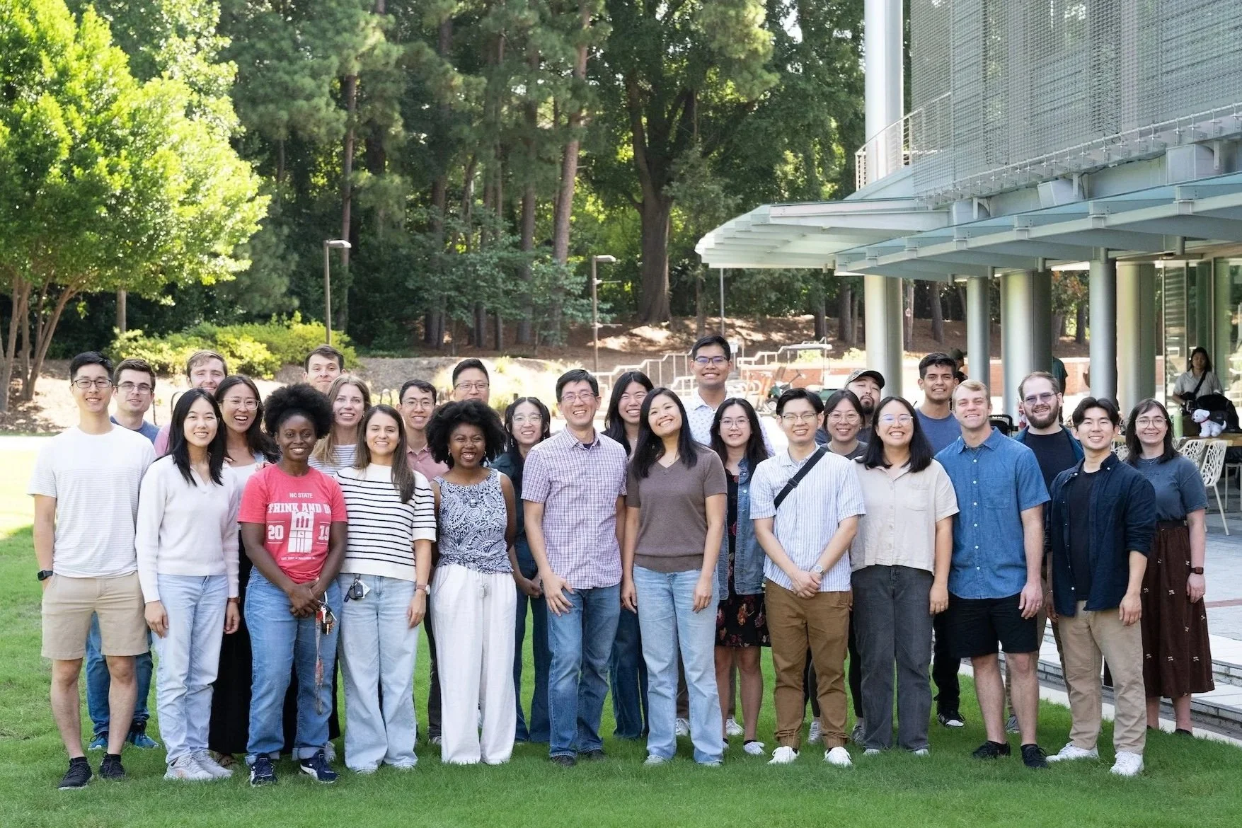 Group of diverse young adults smiling outdoors in front of a building, with trees and a grassy area behind them.