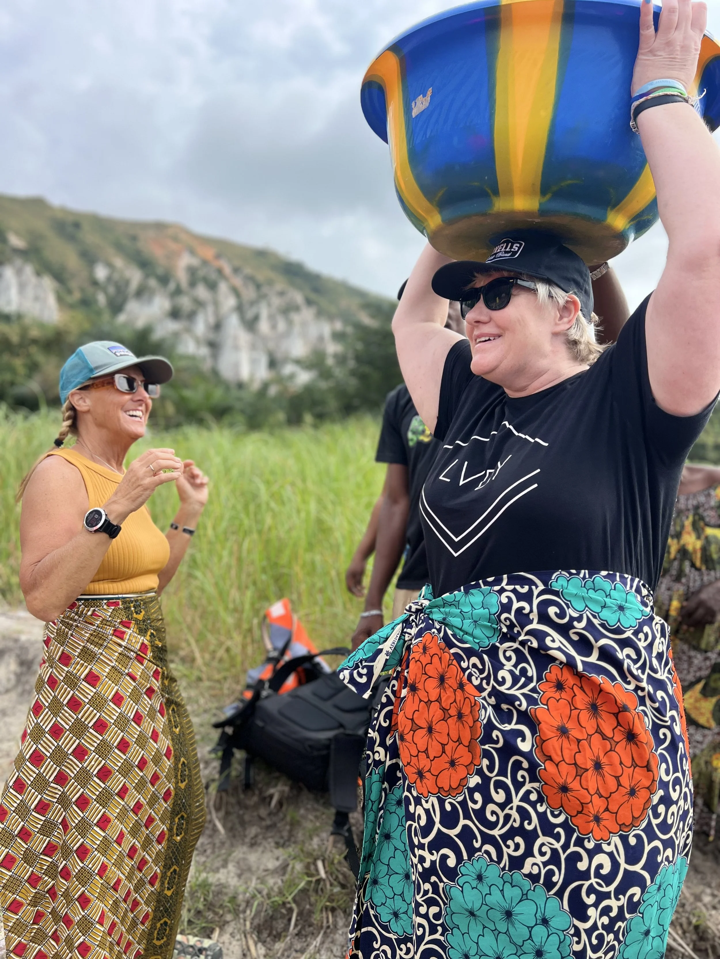 A group of people outdoors, with two women interacting, wearing colorful clothing and sunglasses, with a man balancing a large decorative bowl on his head. The background feature mountains and cloudy sky in the Republic of Congo.