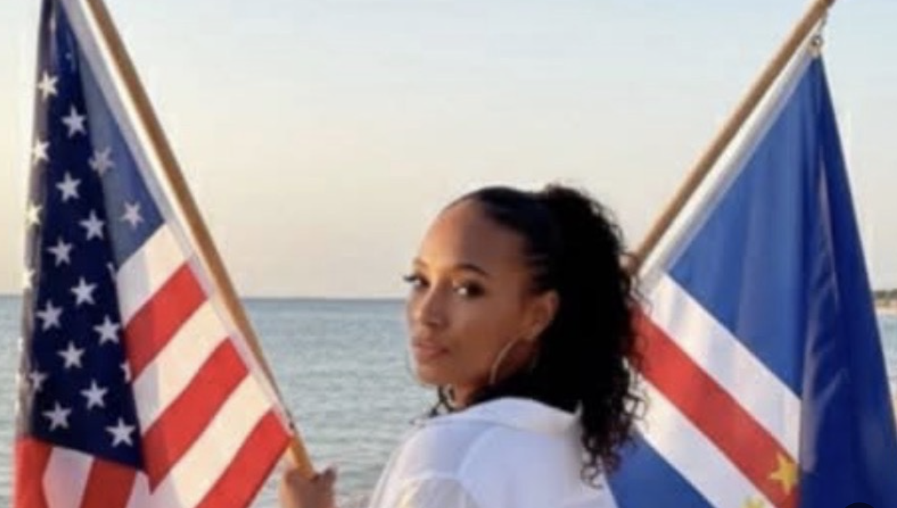 Woman with curly hair holding American and French flags on a beach.