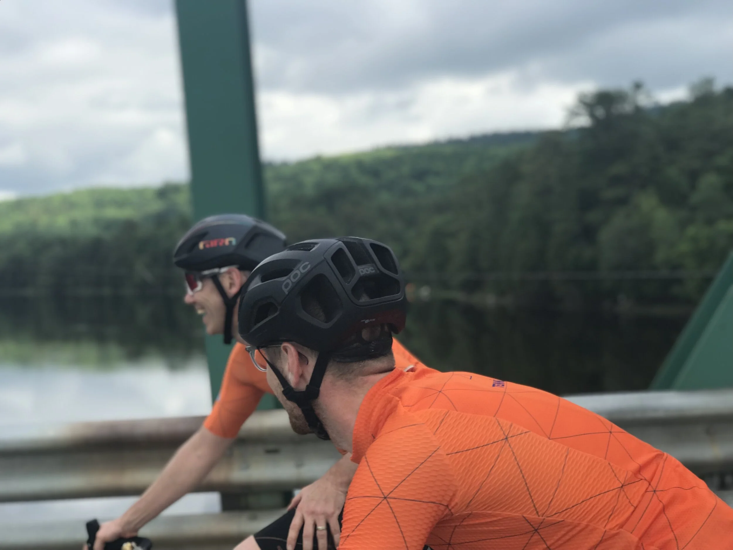 Two cyclists wearing black helmets and orange shirts riding on a bridge with a river and green trees in the background.