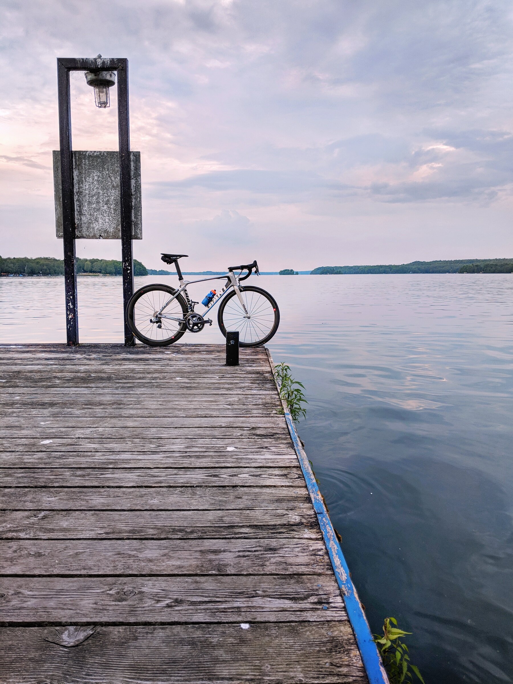 A bicycle parked on a wooden dock by a calm lake with a cloudy sky overhead.