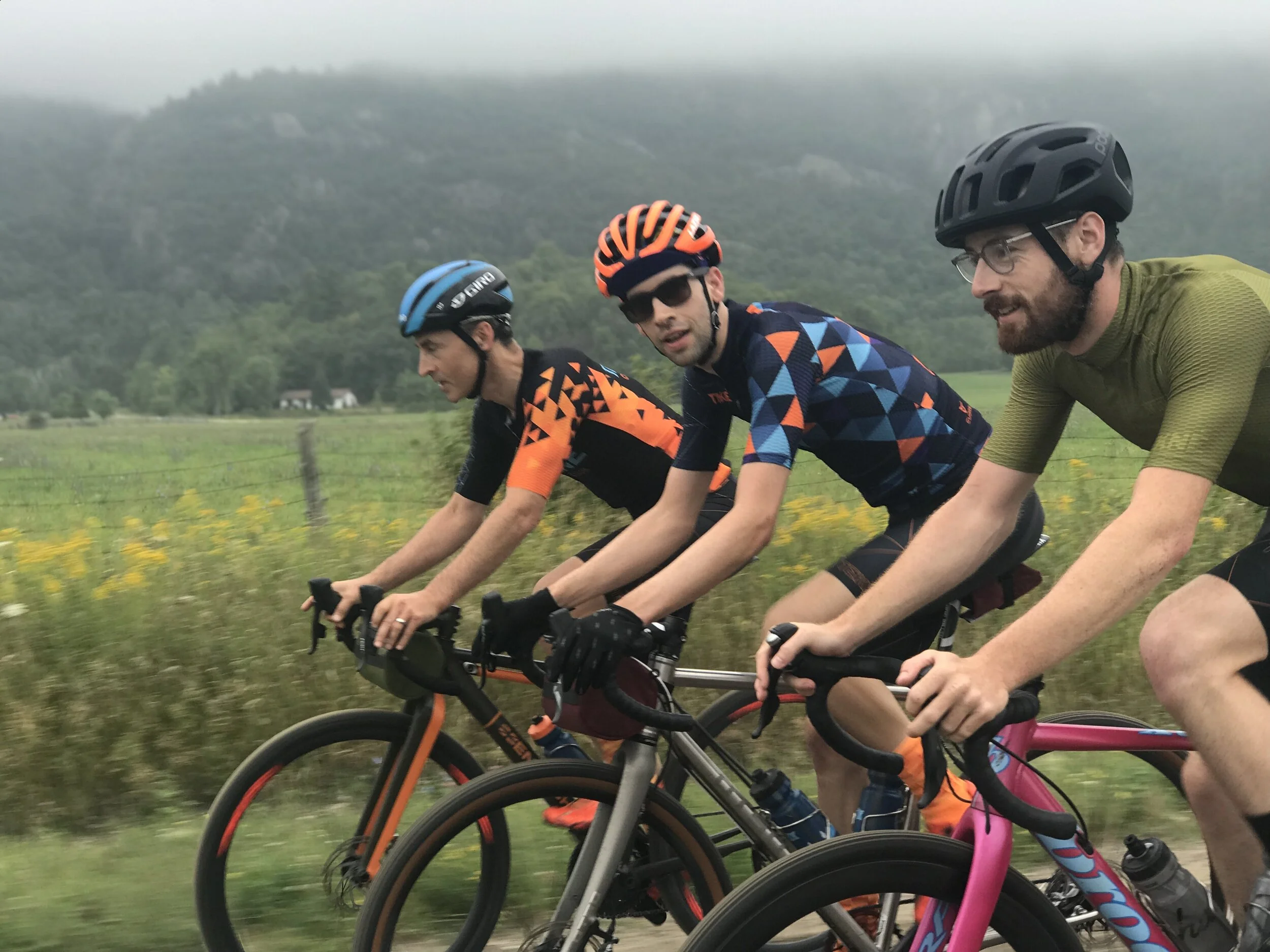 Three men riding bicycles along a rural road with green fields and mountains in the background, wearing helmets and cycling gear.