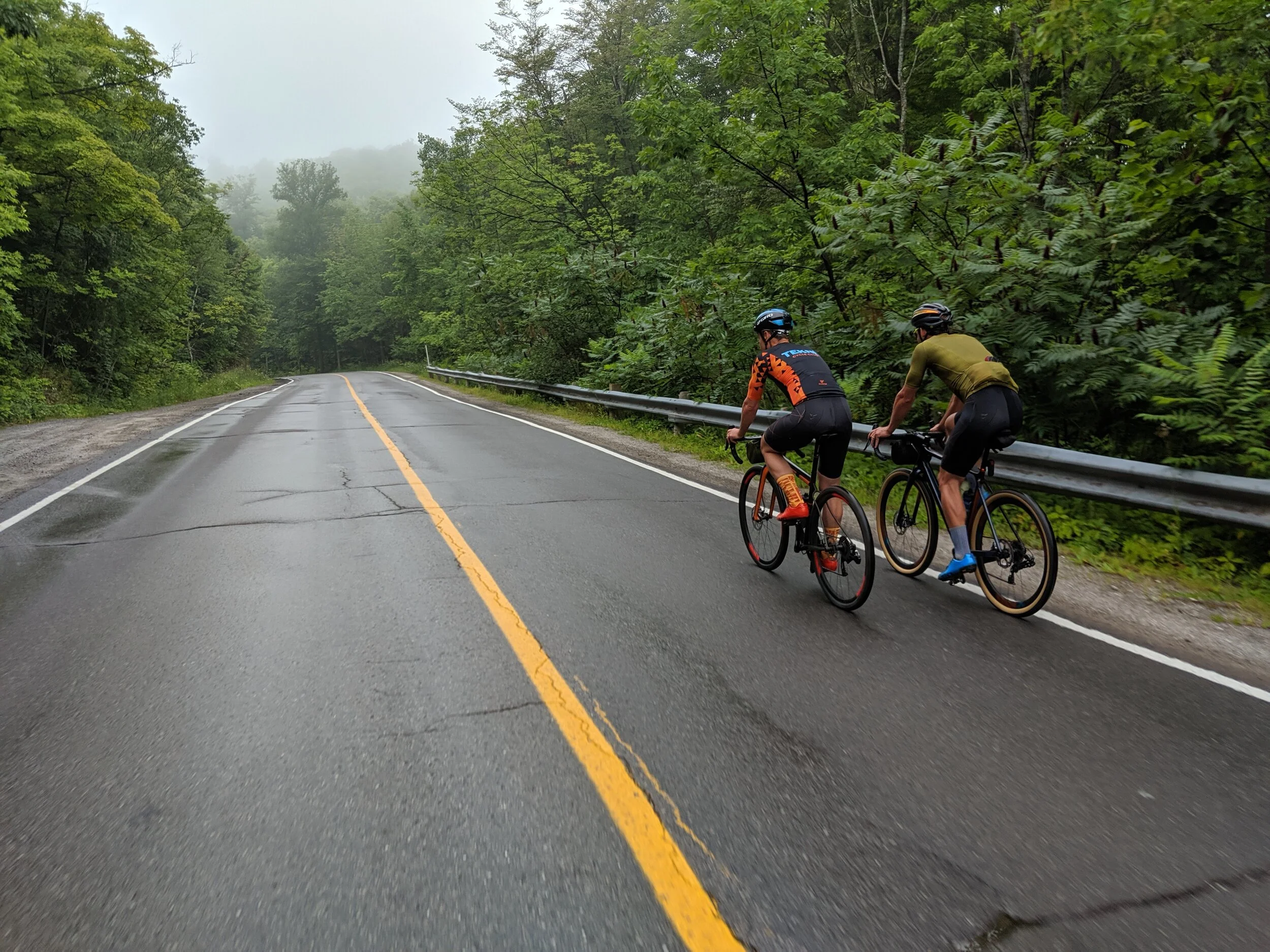 Two cyclists ride on a wet, winding mountain road flanked by lush green trees, with fog in the background.