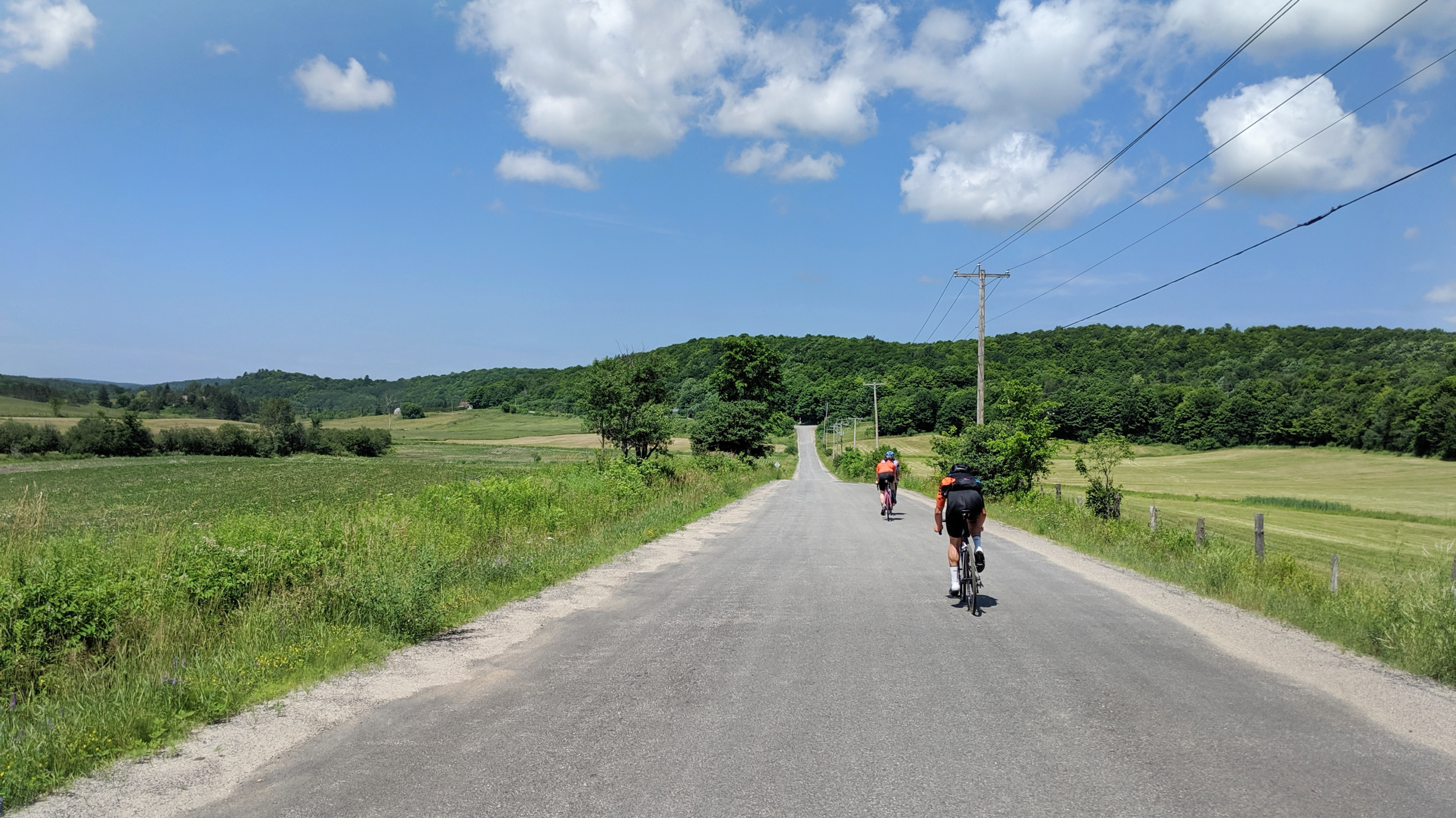 Two cyclists riding on a rural gravel road with green fields and trees on either side under a partly cloudy blue sky.
