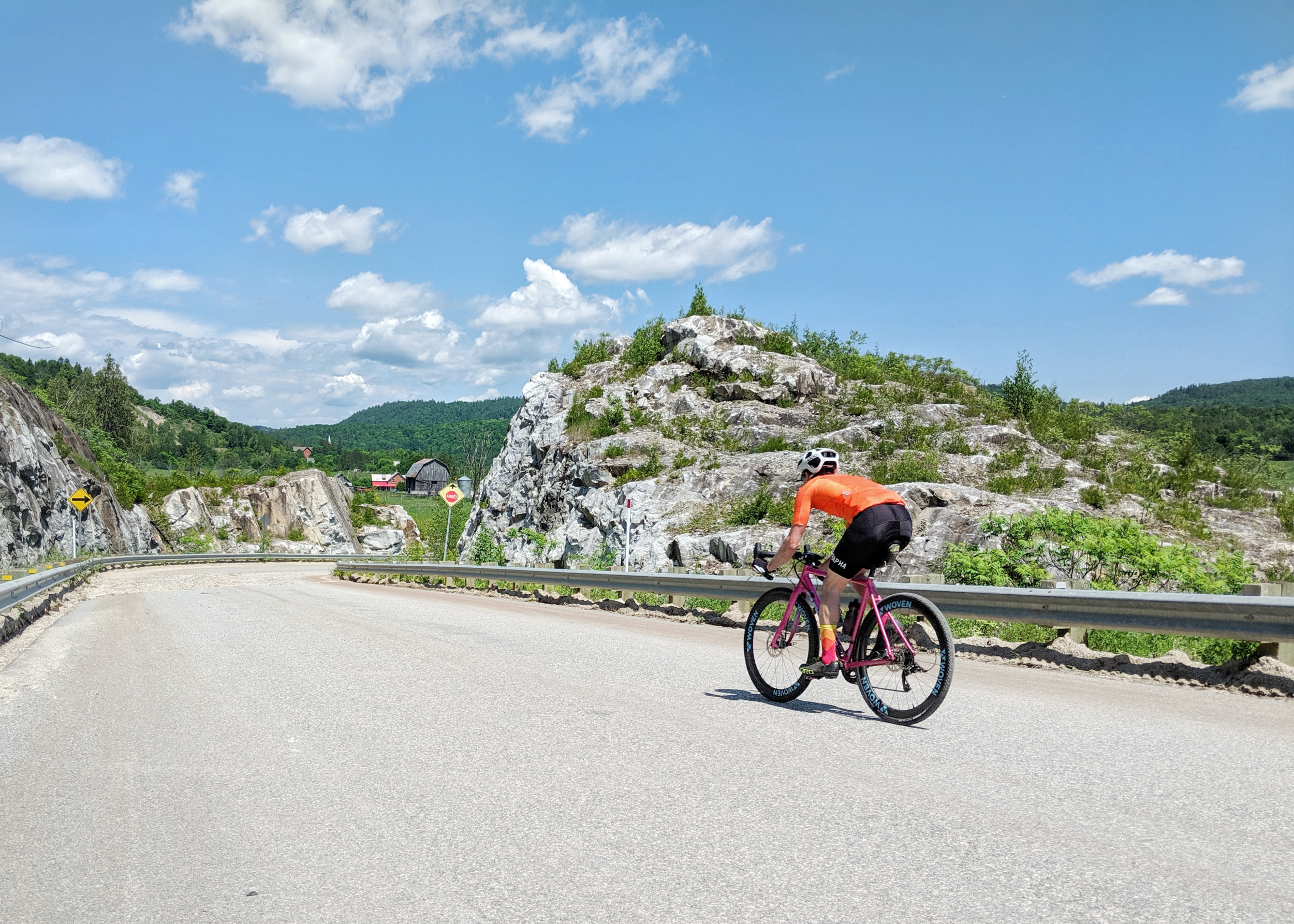 A cyclist in orange shirt and black shorts riding a pink bike on a paved mountain road with rocky cliffs, green trees, and a blue sky with white clouds in the background.