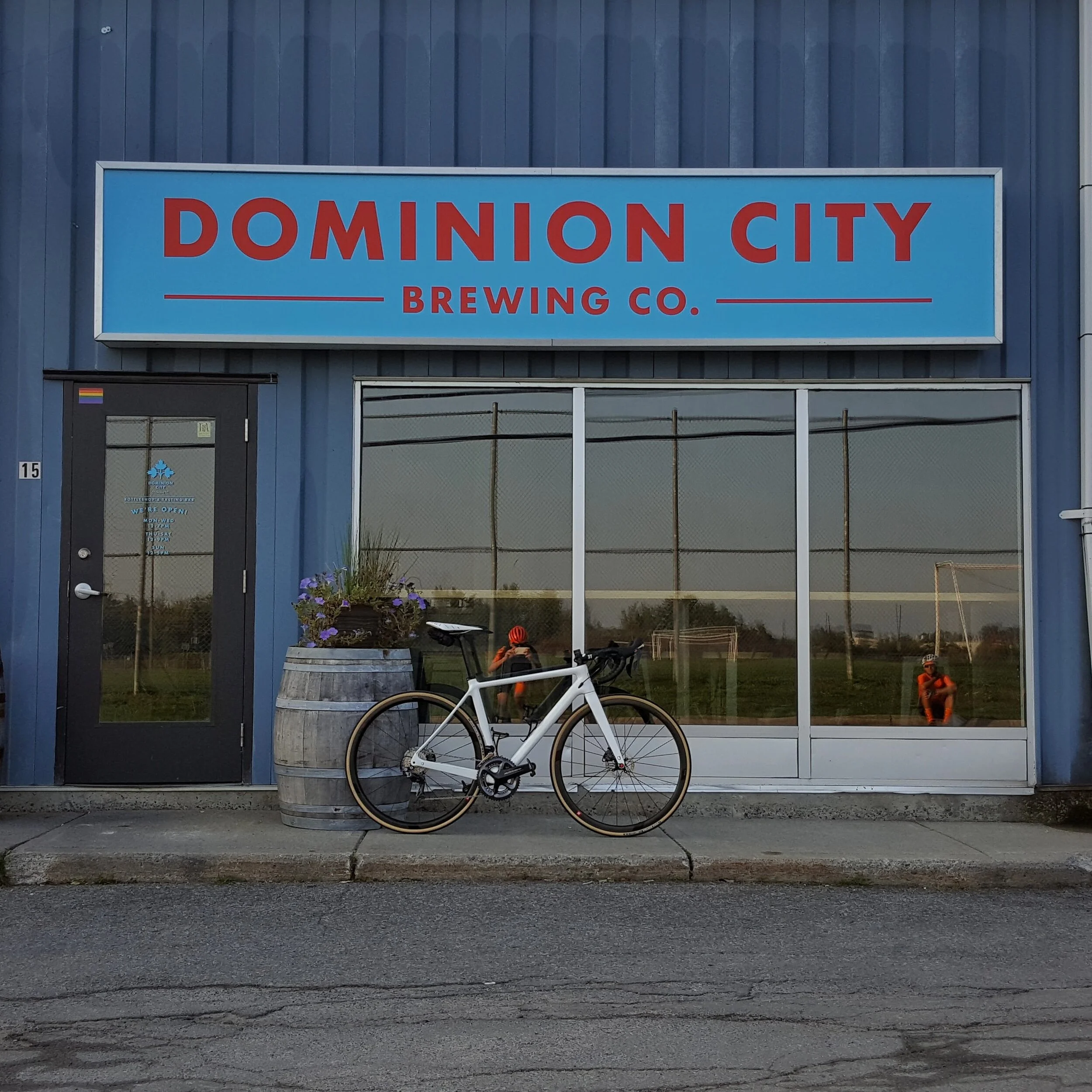 Exterior of Dominion City Brewing Co. with a bicycle parked in front, a large window, and a barrel with flowers outside.