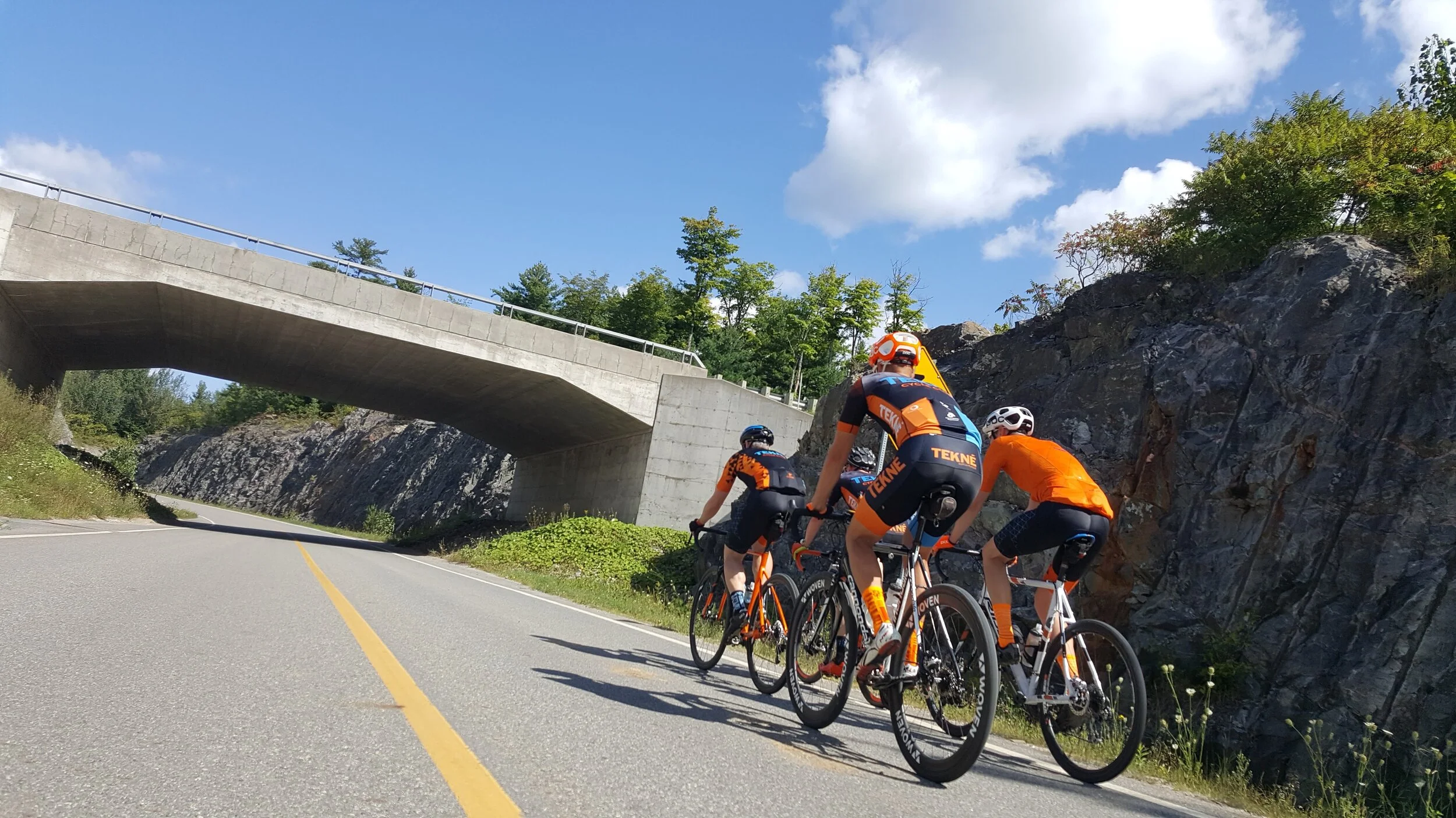 Group of four cyclists riding uphill on a mountain road beneath a concrete bridge with a rocky cliff on one side and green trees on top, under a sunny sky with some clouds.