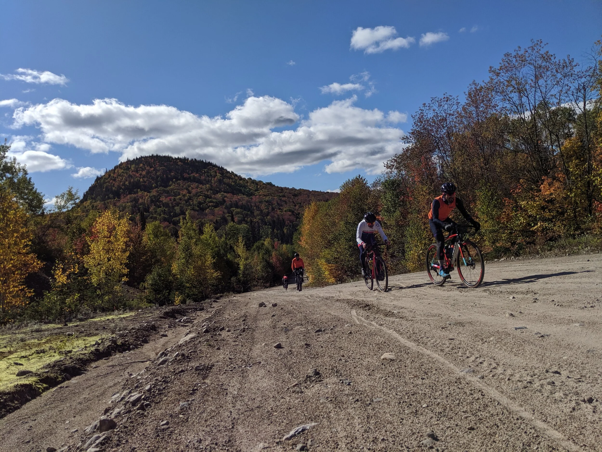 Four cyclists riding on a dirt trail surrounded by trees with fall foliage, under a partly cloudy blue sky, with a mountain in the background.