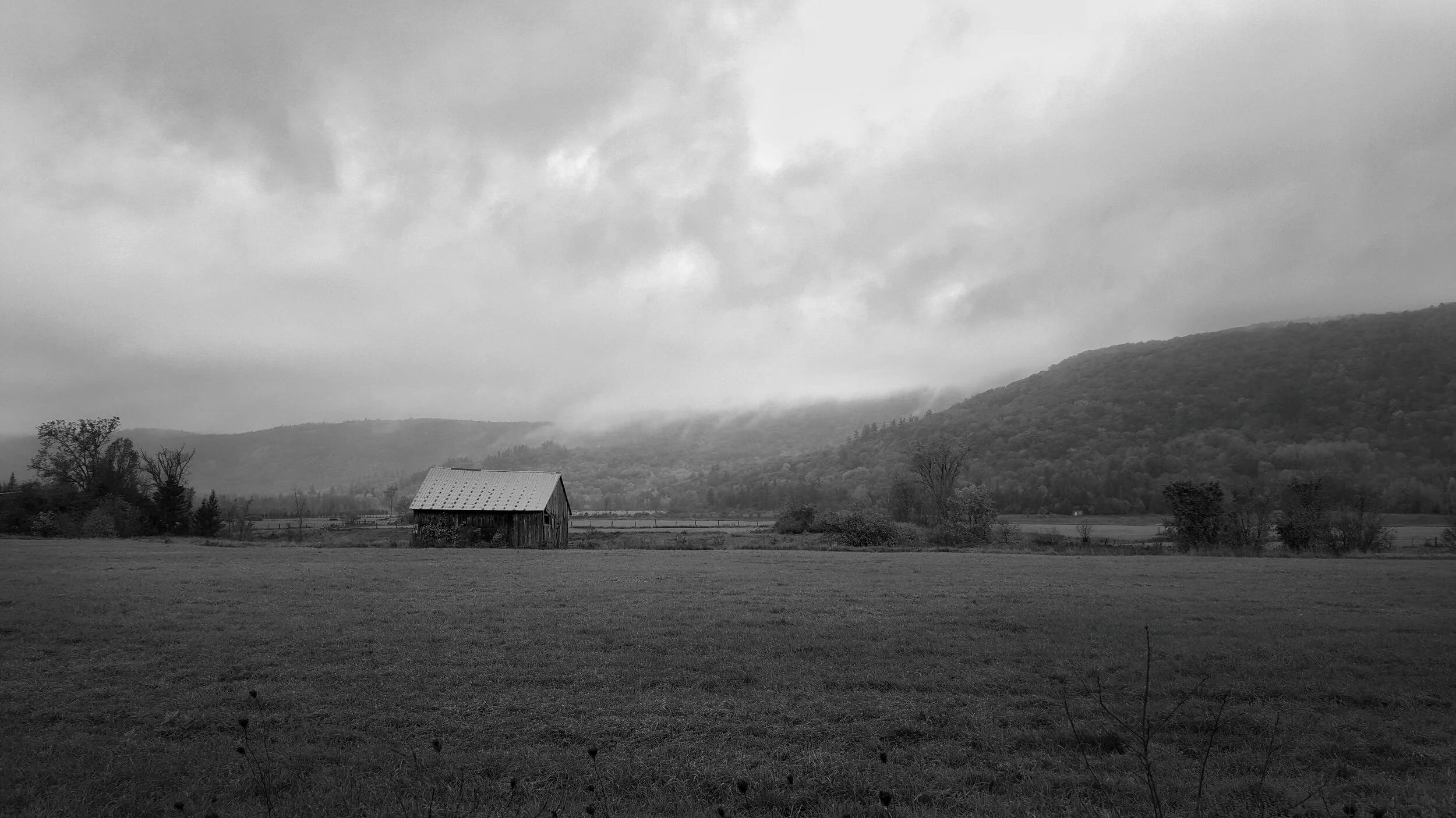 A black and white photo of a rural landscape with a small wooden barn in the foreground, open grass fields, sparse trees, and mountains in the background under a cloudy sky.
